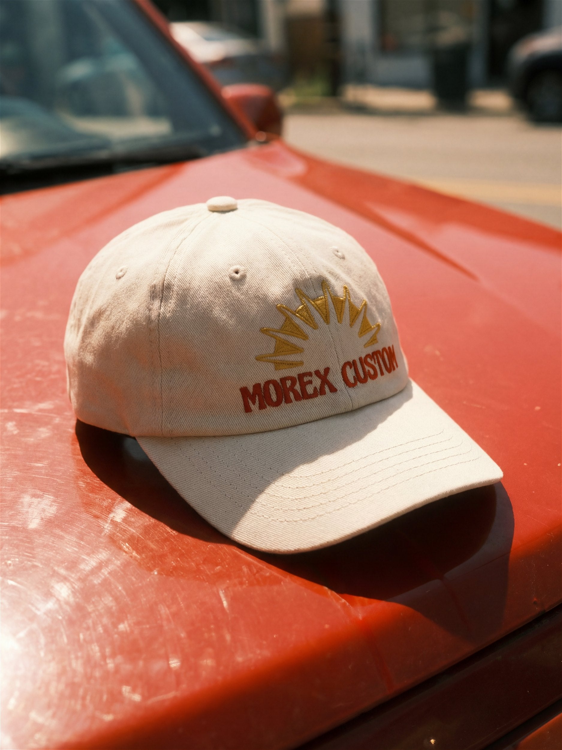 Casquette Morex Custom beige avec broderie logo soleil rouge et jaune, présentée sur le capot d'une voiture de collection rouge.
