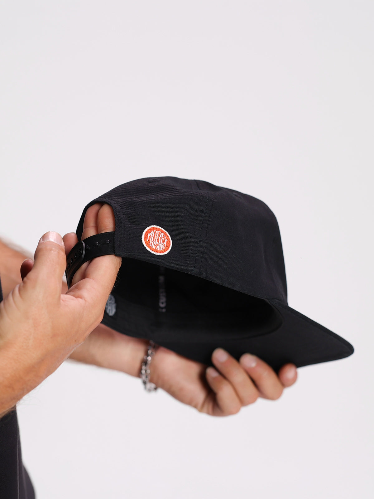 Gros plan sur l'arrière d'une casquette noire Morex Custom avec un patch logo circulaire rouge, tenue par une personne dans un style lifestyle moto.