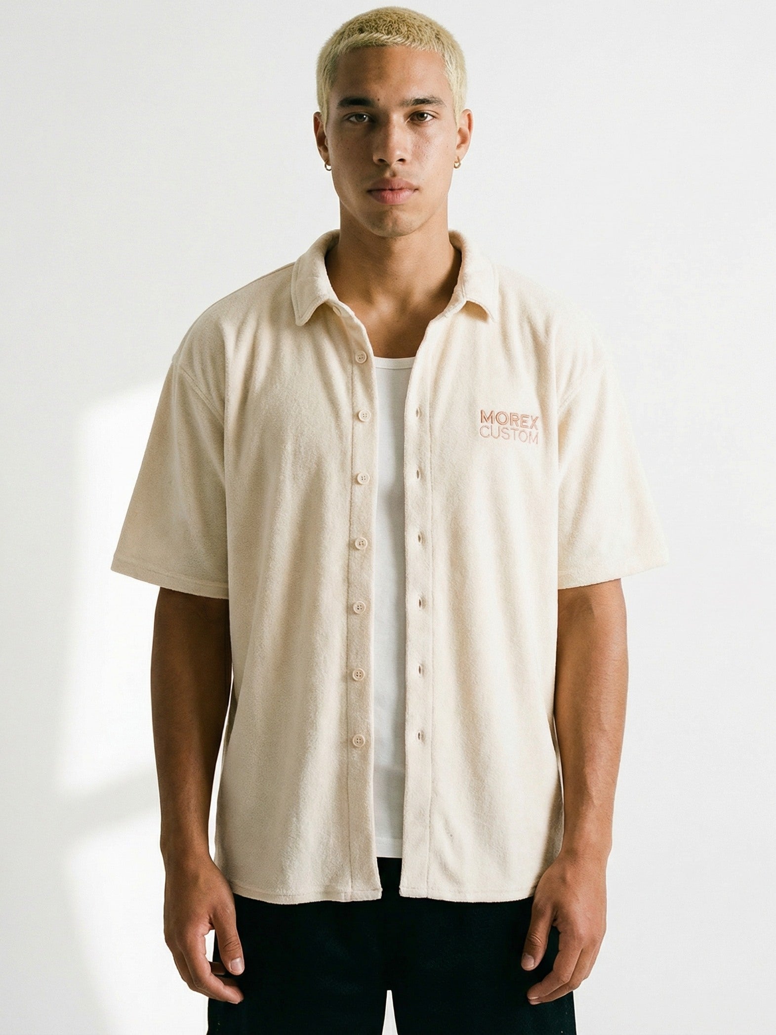 Un homme aux cheveux blonds courts portant une chemise beige à manches courtes de la marque Morex Custom sur un débardeur blanc.