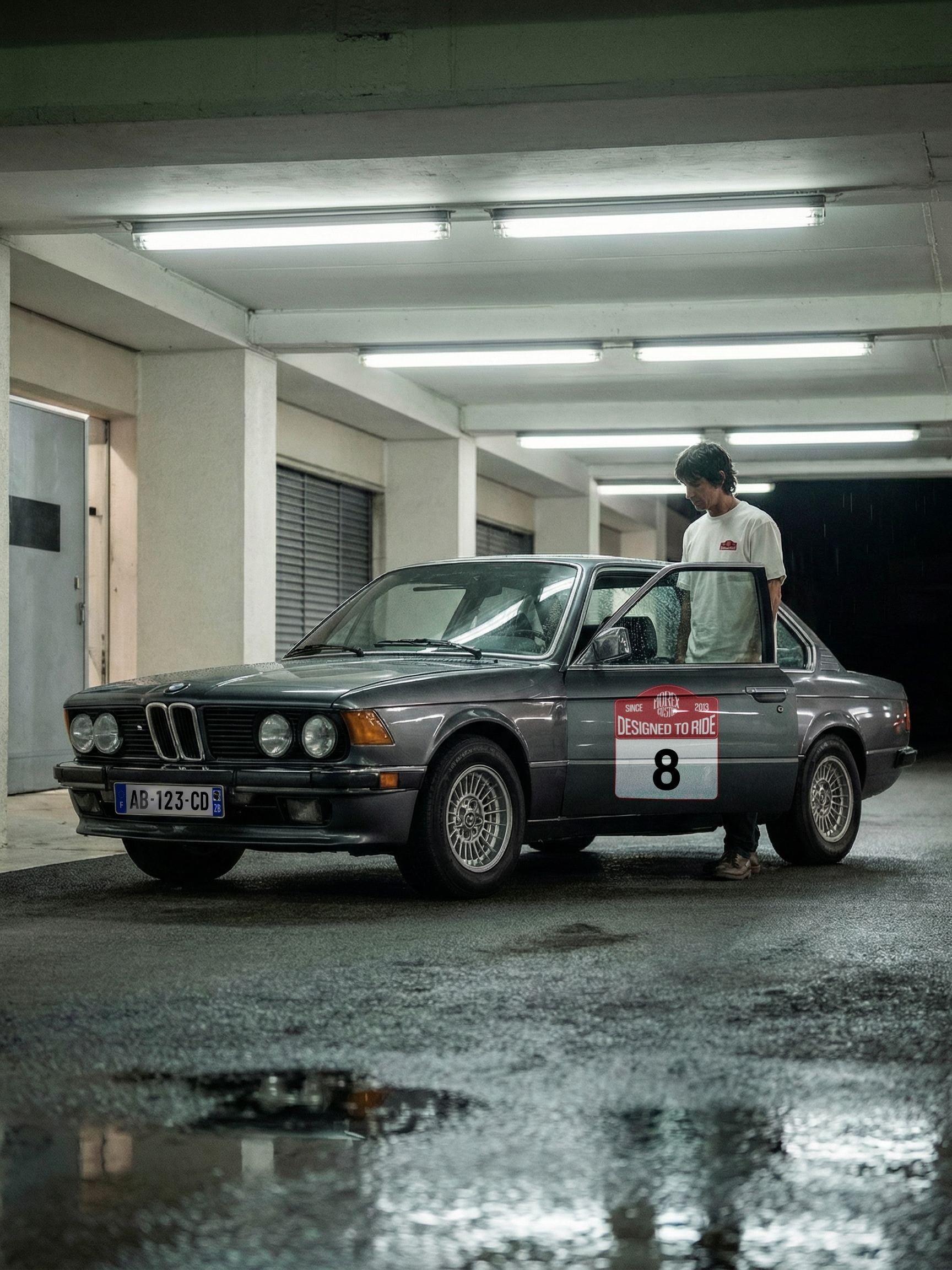 Une BMW E21 classique de couleur anthracite garée dans un parking souterrain humide, avec un homme debout à côté de la portière ouverte portant un sticker Morex Custom 'Designed to Ride' numéro 8.