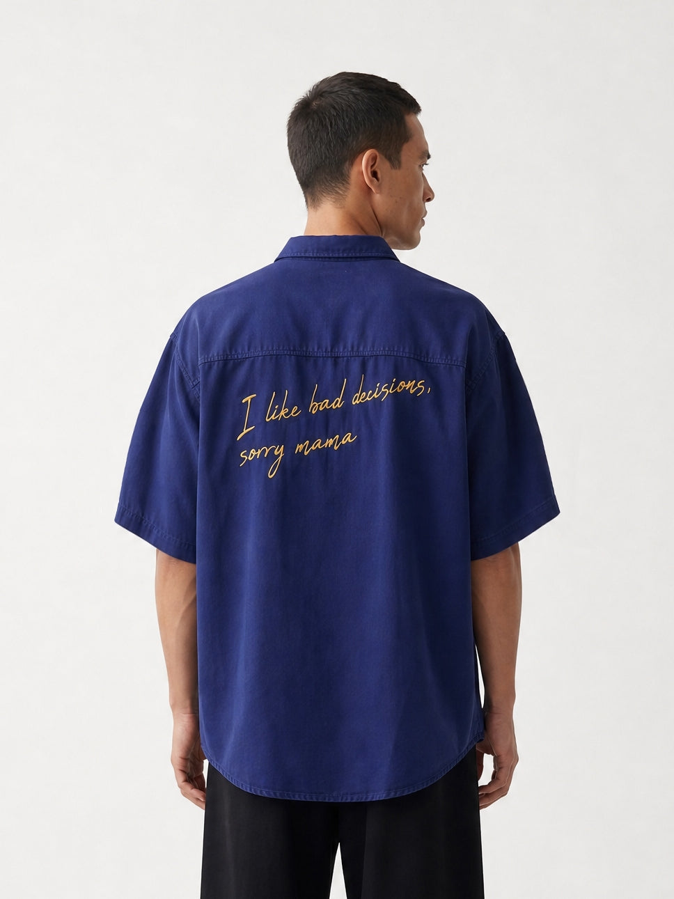 Vue arrière d'un homme portant une chemise à manches courtes bleu marine Morex Custom avec une broderie jaune cursive au dos, associée à un pantalon large noir.