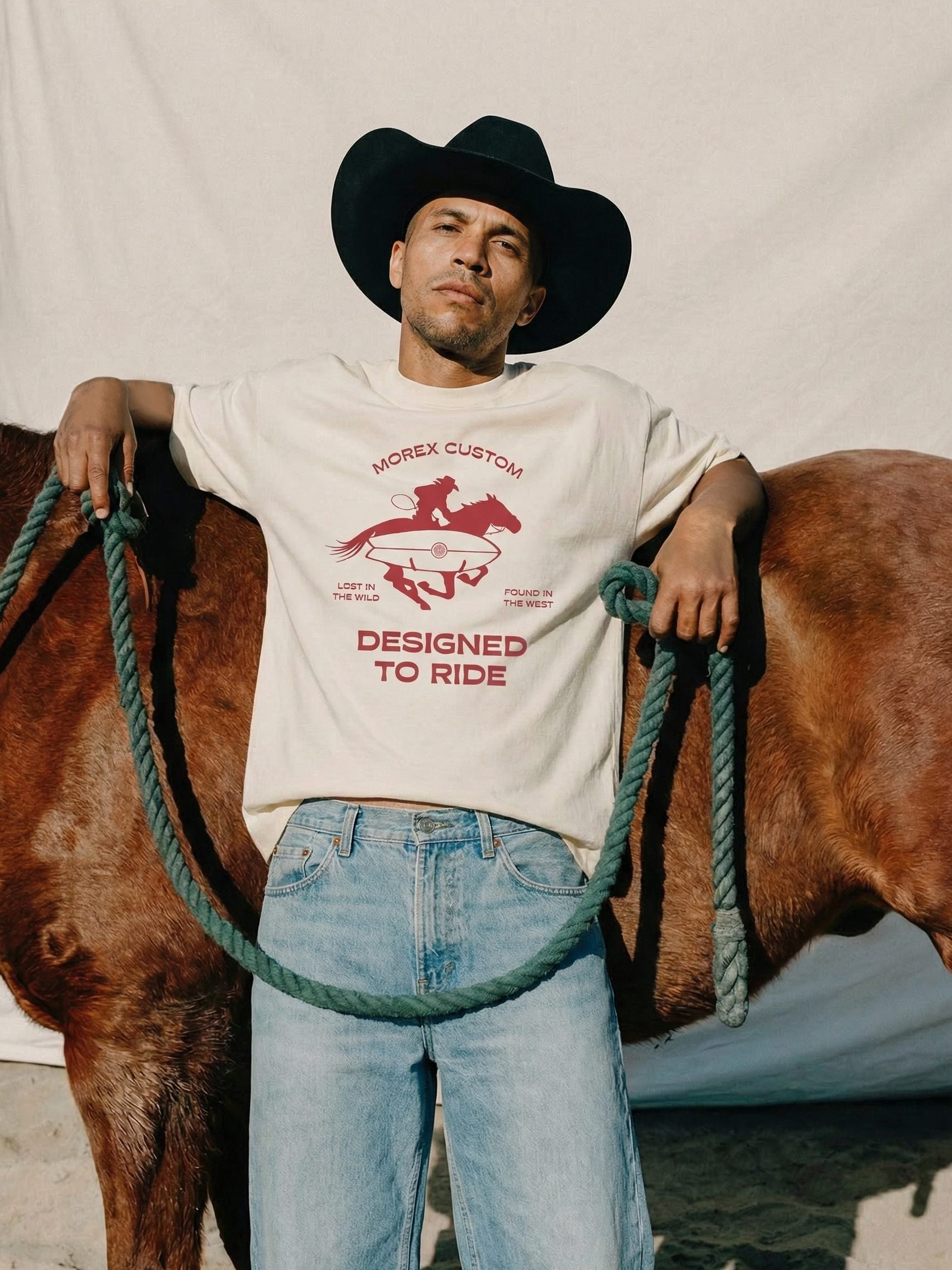 A man wearing a black cowboy hat and a Morex Custom 'Designed to Ride' cream t-shirt, leaning against a brown horse while holding a green rope.