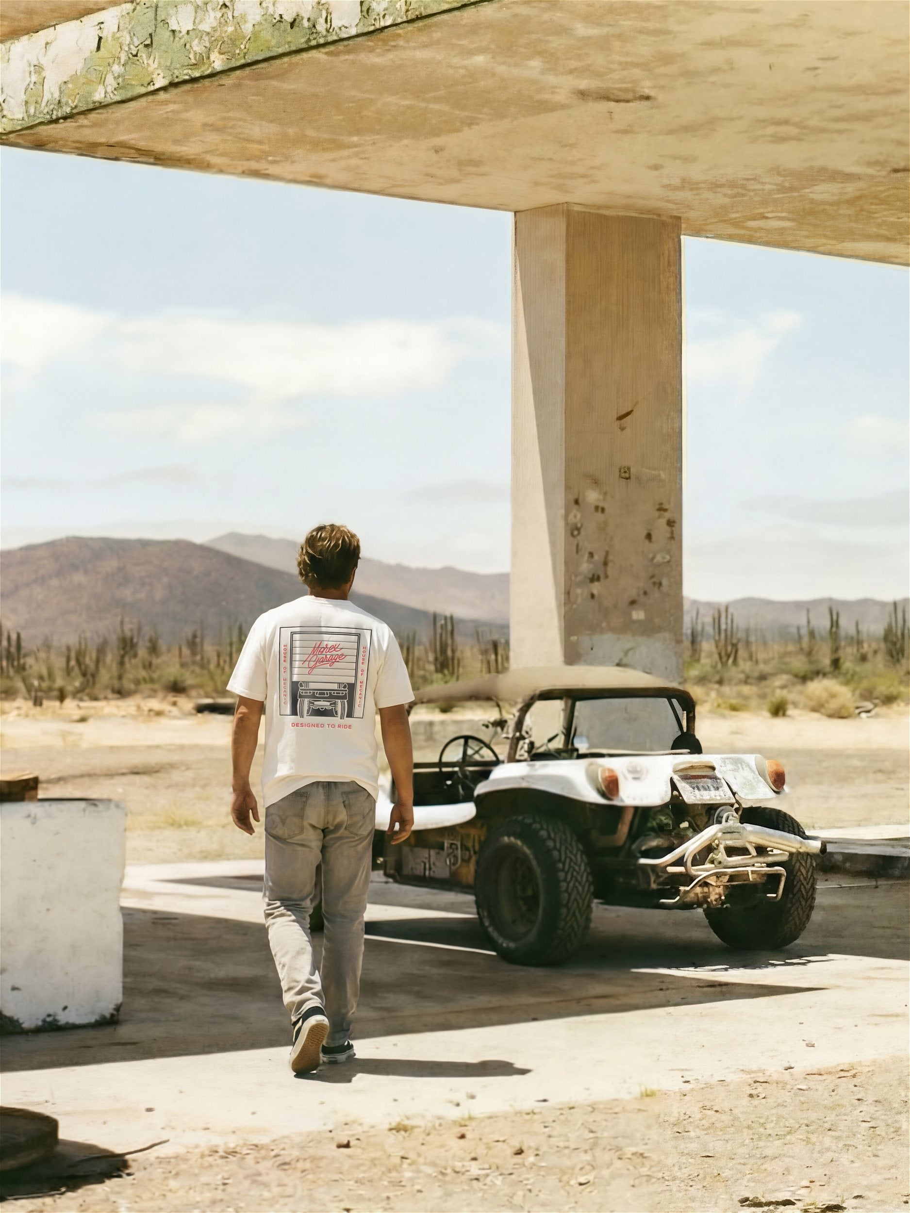 Photographie lifestyle Morex Custom montrant un homme de dos marchant vers un buggy blanc dans un paysage désertique aride sous un auvent vintage.