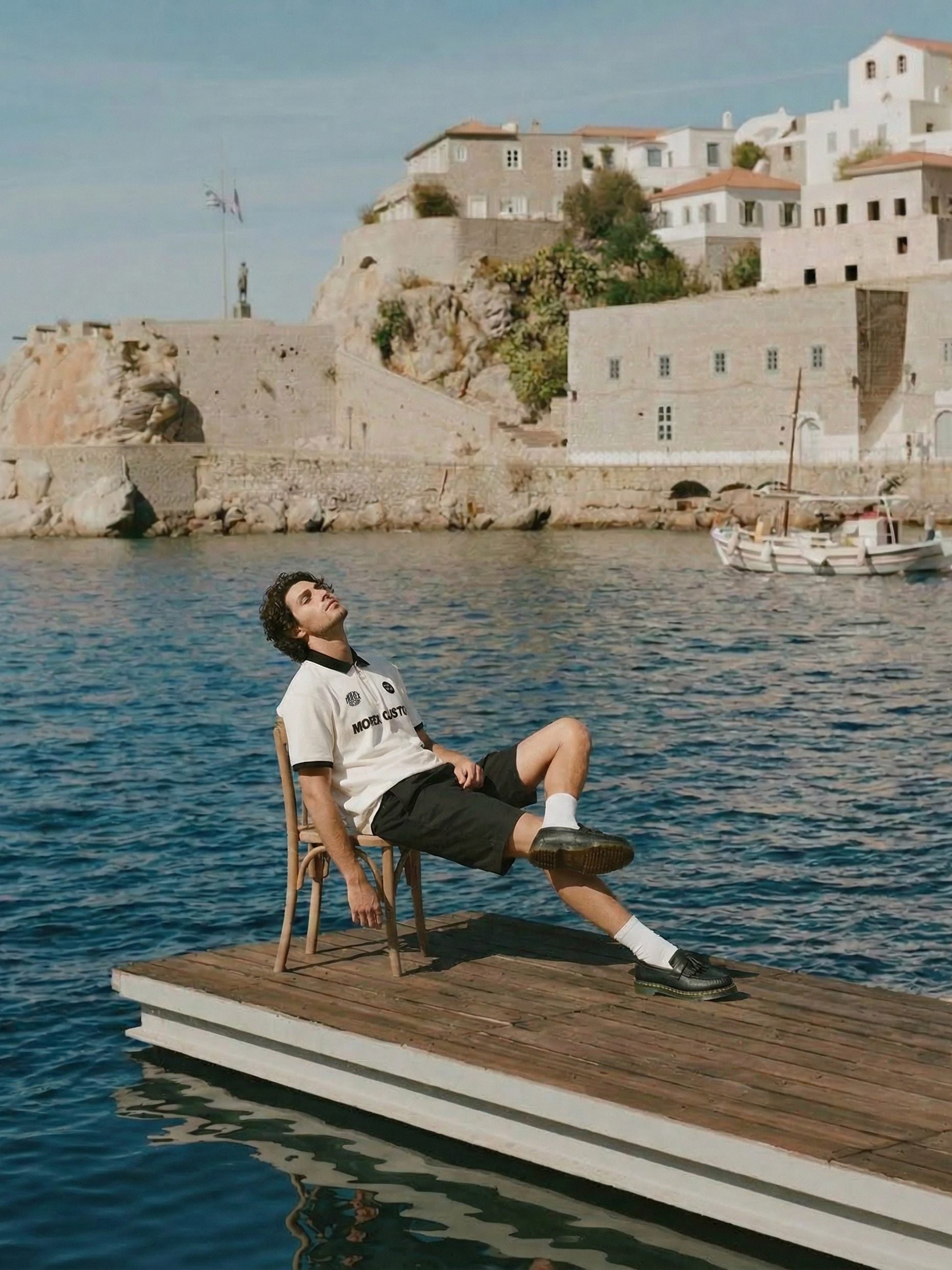 Homme portant un polo blanc Morex Custom et des mocassins, assis sur un quai en bois devant un village côtier méditerranéen.