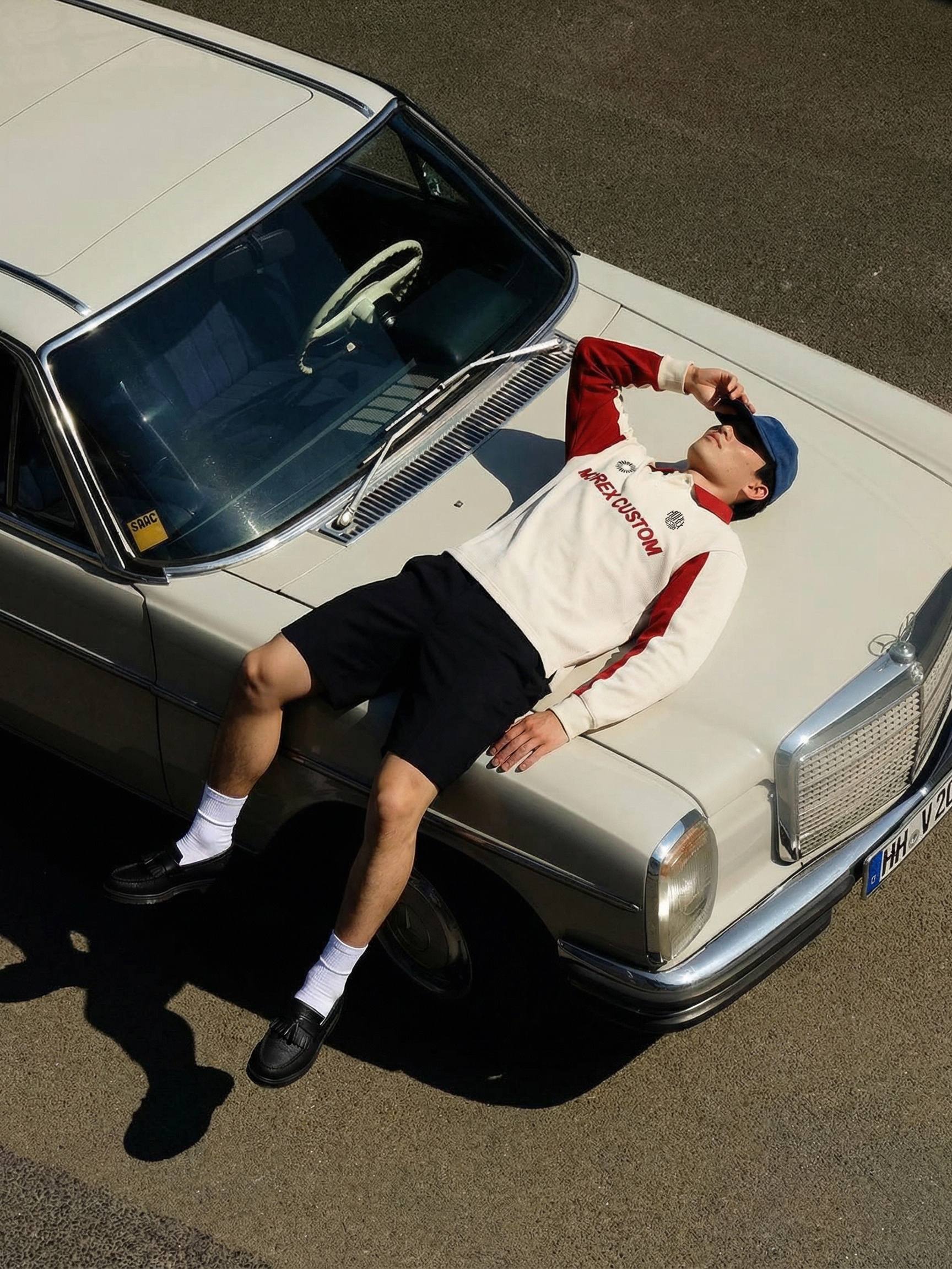 Un homme portant un sweat-shirt Morex Custom blanc et rouge, allongé sur le capot d'une voiture Mercedes-Benz vintage blanche pour un look lifestyle décontracté.