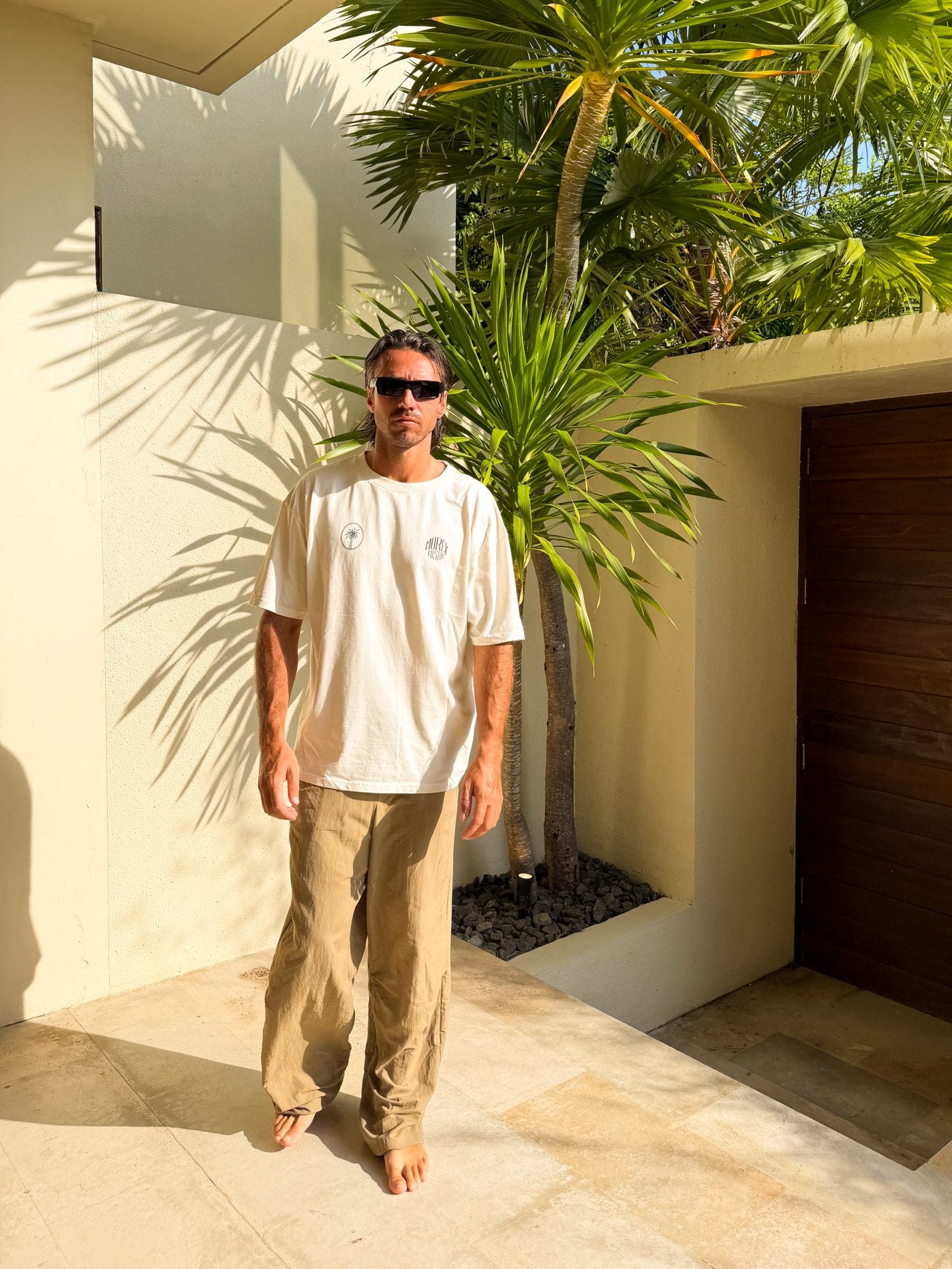 Homme portant un t-shirt blanc et un pantalon beige de la collection Morex Custom, posant dans un décor tropical ensoleillé avec des palmiers.