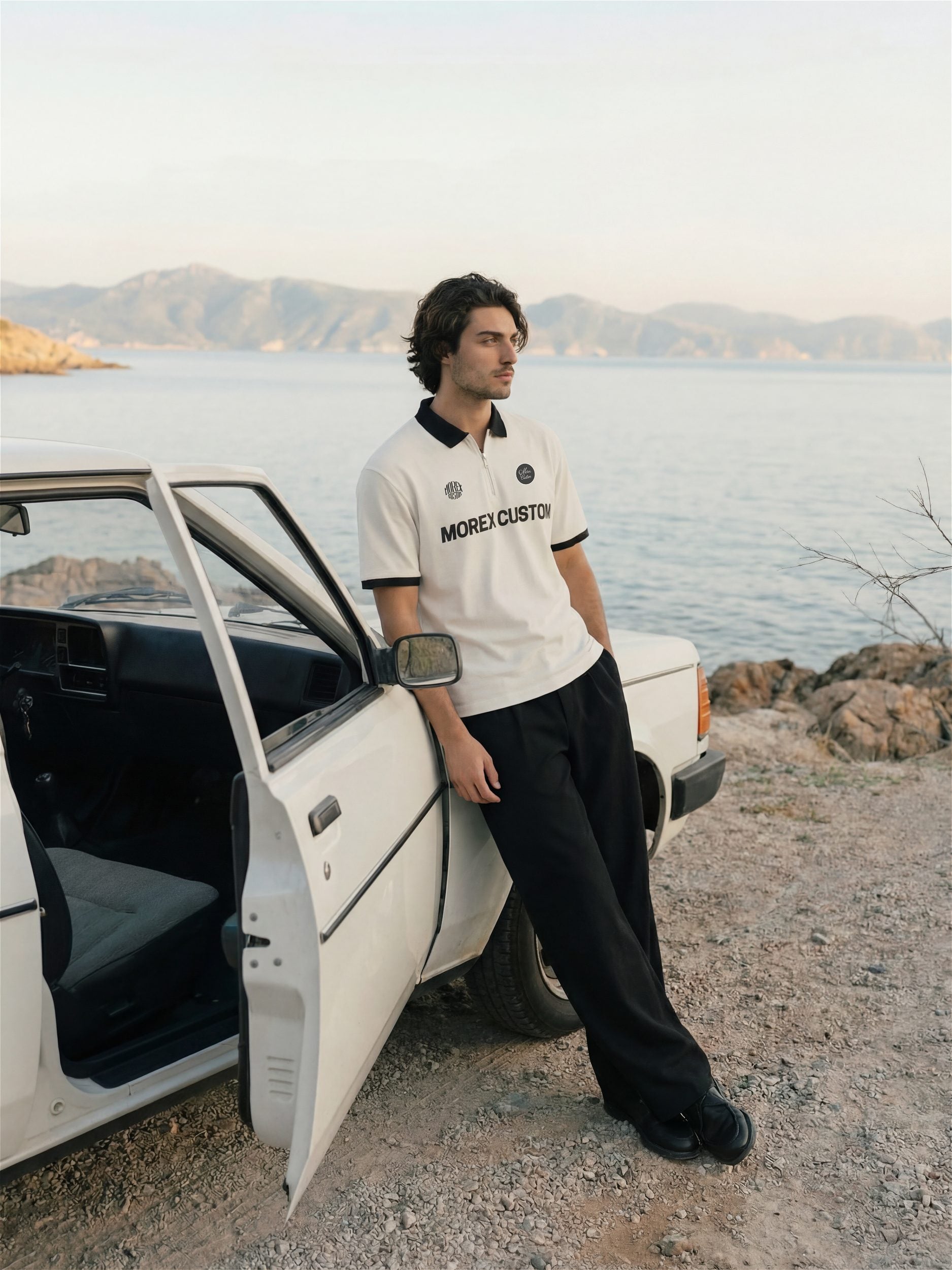 Un homme portant un polo Morex Custom blanc avec détails noirs, s'appuyant sur une voiture vintage blanche sur une route côtière au coucher du soleil.