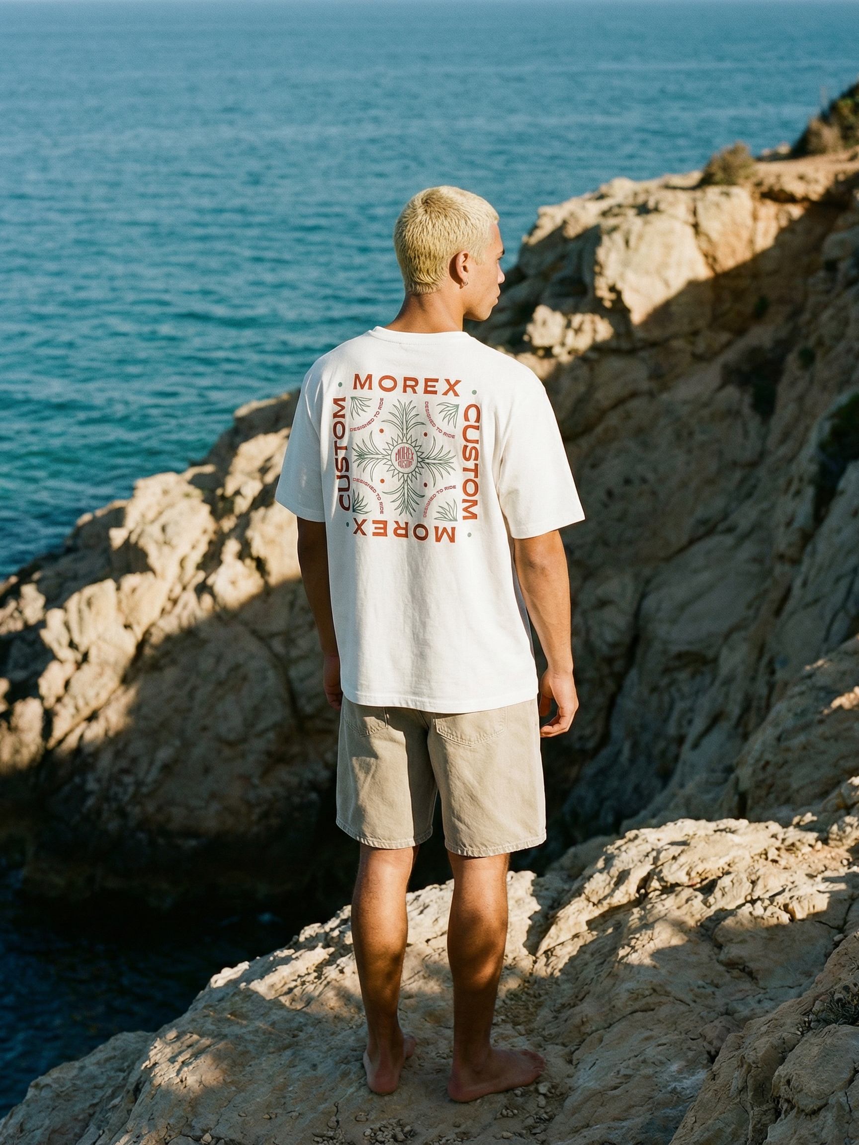 Homme de dos portant un t-shirt blanc Morex Custom avec un large graphique carré au dos, debout sur une falaise rocheuse face à l'océan.