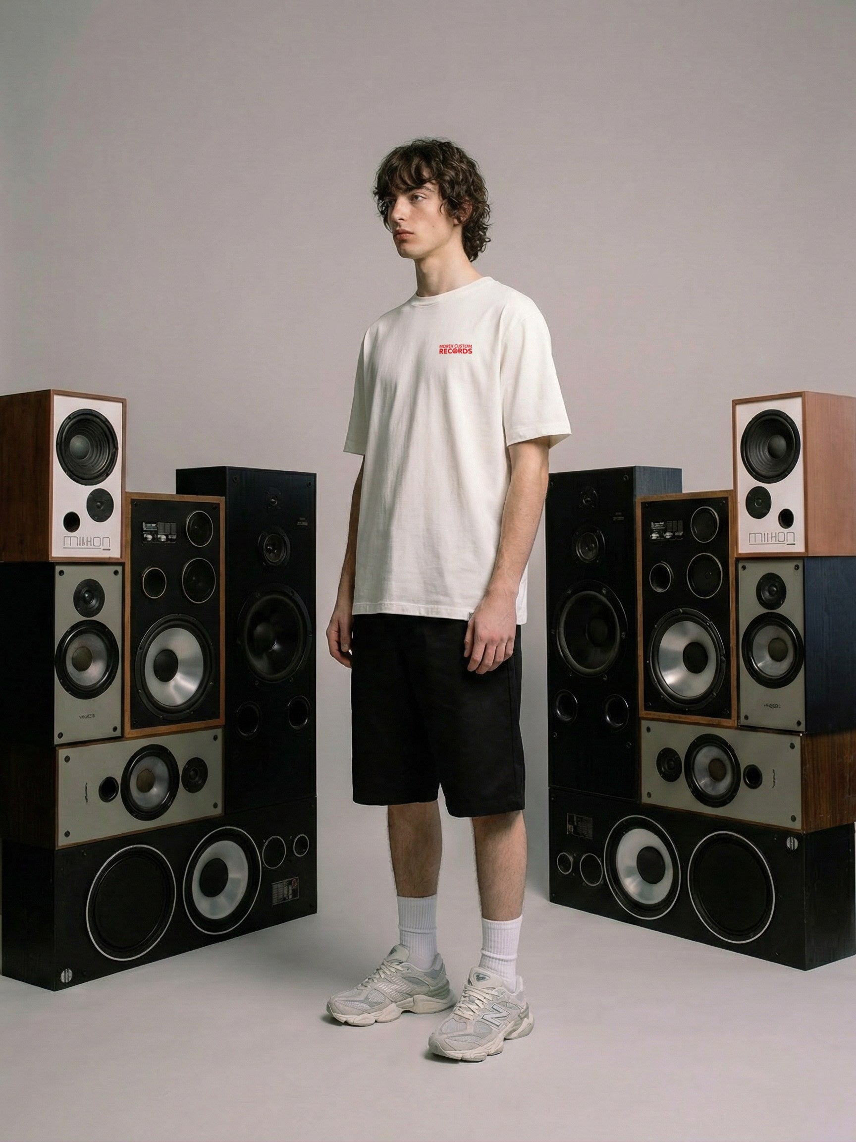 Un homme posant avec un t-shirt blanc Morex Custom à logo rouge, entouré de deux colonnes d'enceintes audio vintage.