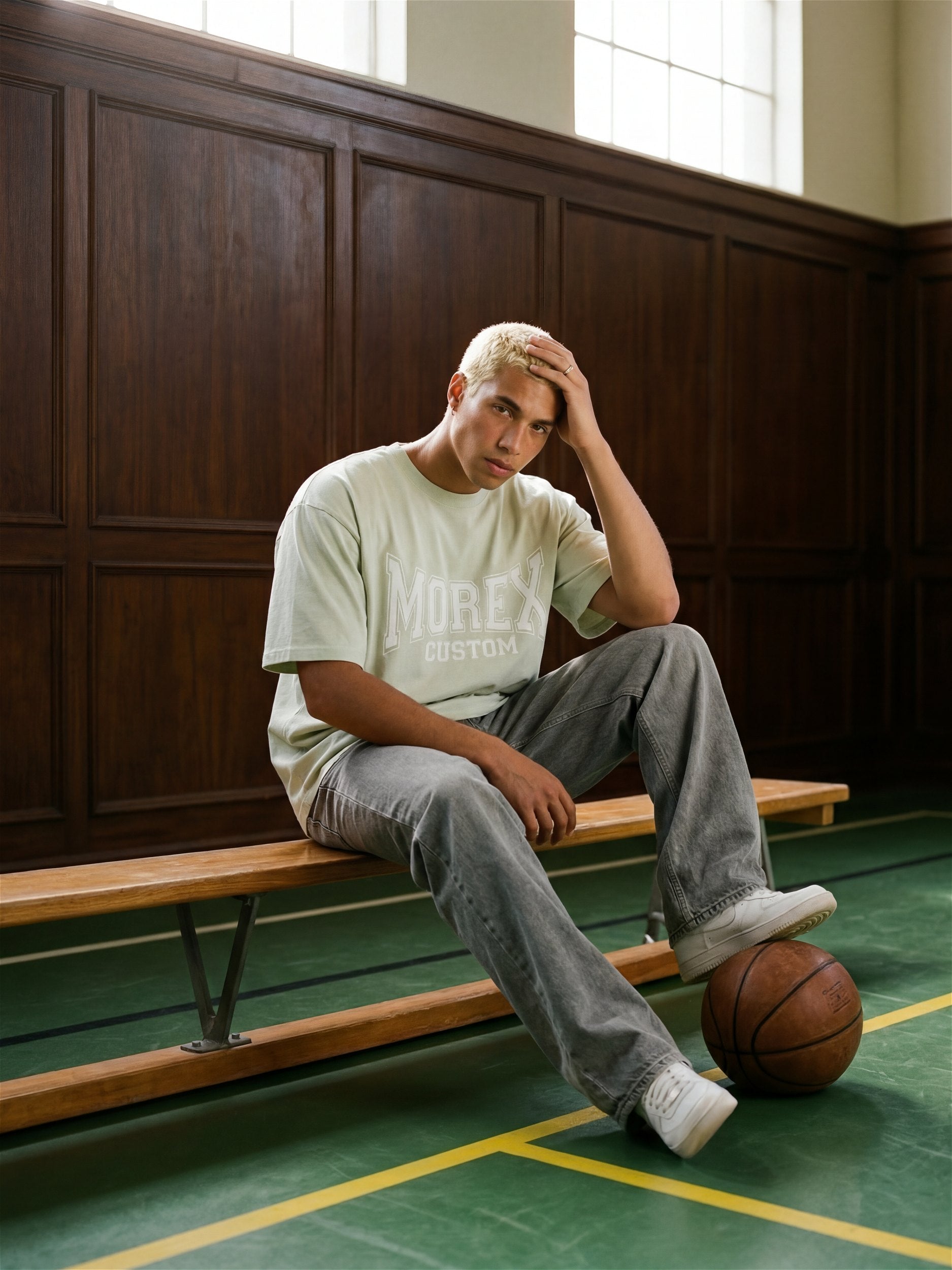 Un homme portant un t-shirt Morex Custom de style varsity couleur vert sauge, assis sur un banc de gymnase avec un ballon de basket.