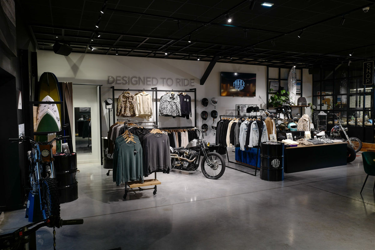 Vue intérieure du magasin Morex Custom avec une sélection de vêtements de prêt-à-porter, d'accessoires moto et une moto customisée exposée.
