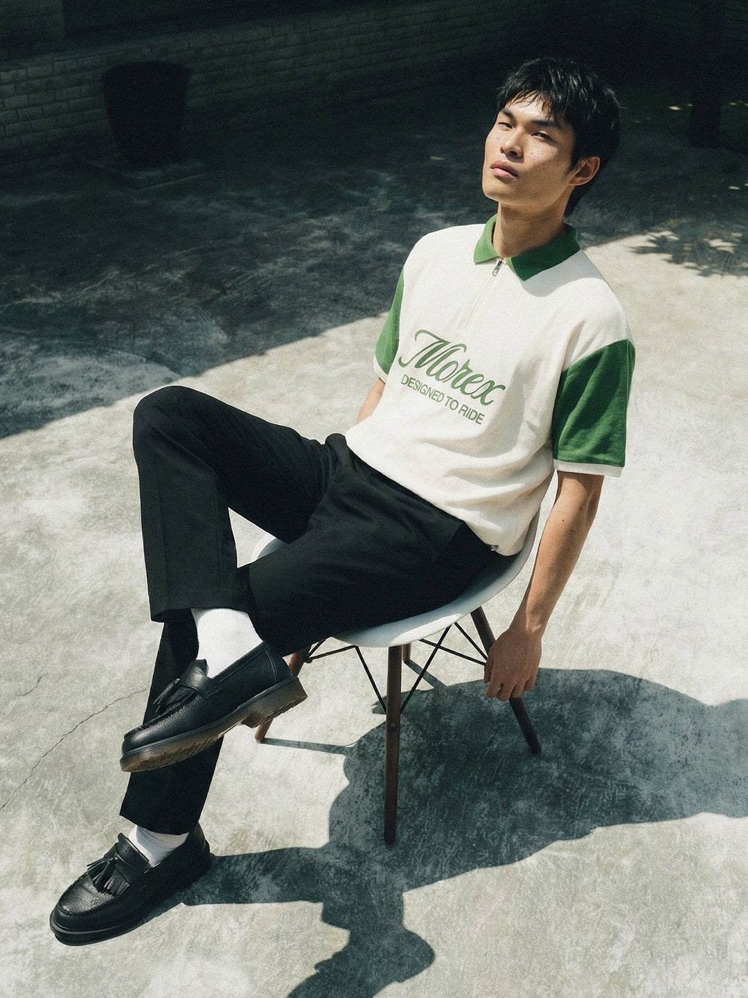 Homme assis en extérieur portant un polo Morex Custom bicolore blanc et vert avec logo brodé, pantalon noir et mocassins, style lifestyle urbain.
