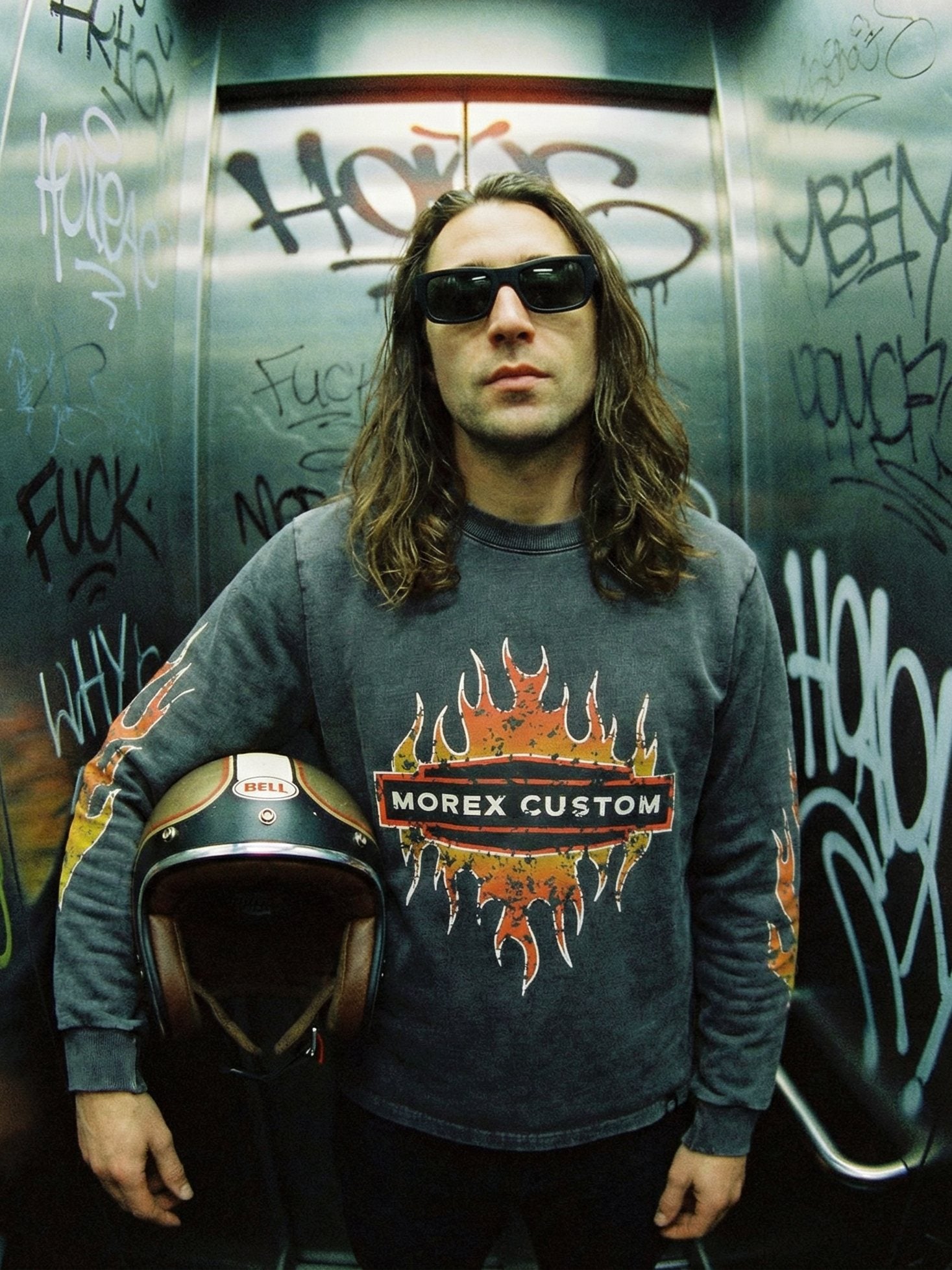 Homme arborant un look lifestyle motard avec un sweat Morex Custom et un casque de moto dans un décor urbain tagué.