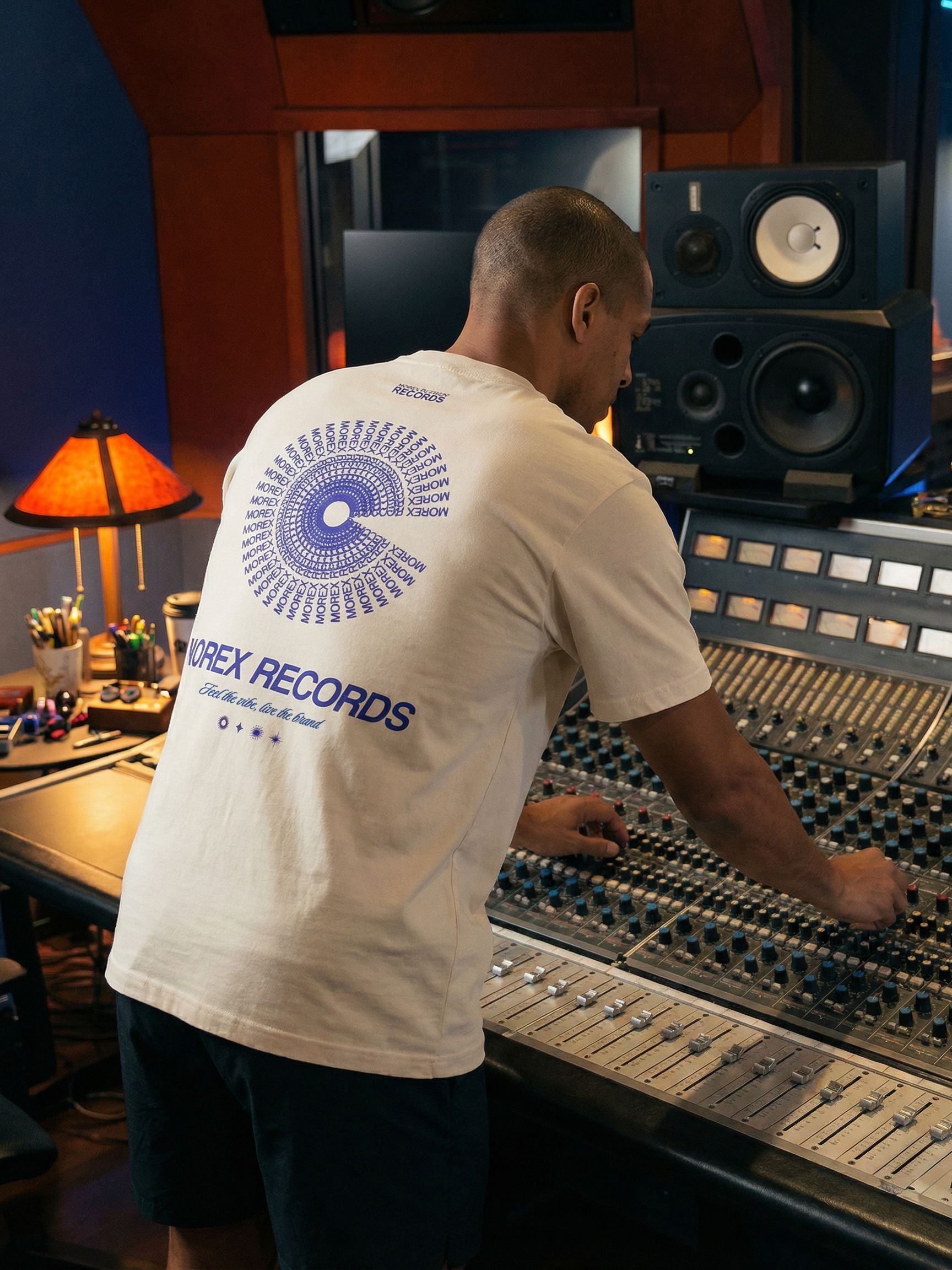 Homme de dos portant un t-shirt beige Morex Records devant une console de mixage dans un studio d'enregistrement.
