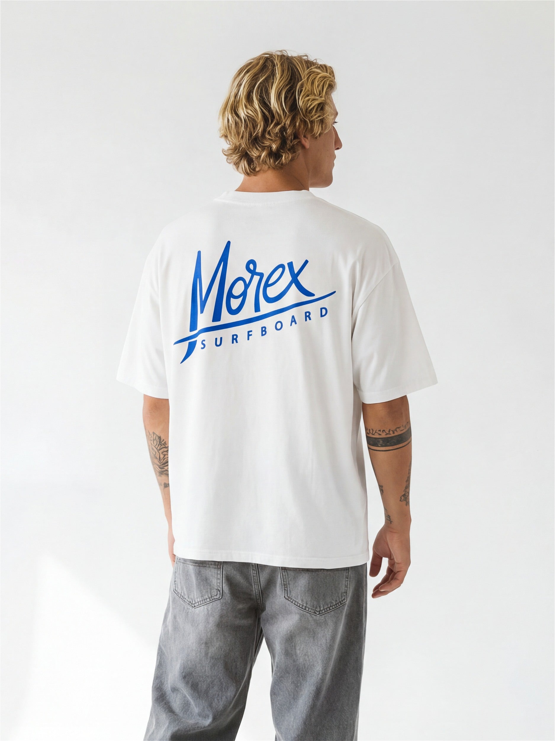 Homme de dos portant un t-shirt blanc Morex Surfboard avec un logo graphique bleu et un jean large gris délavé.