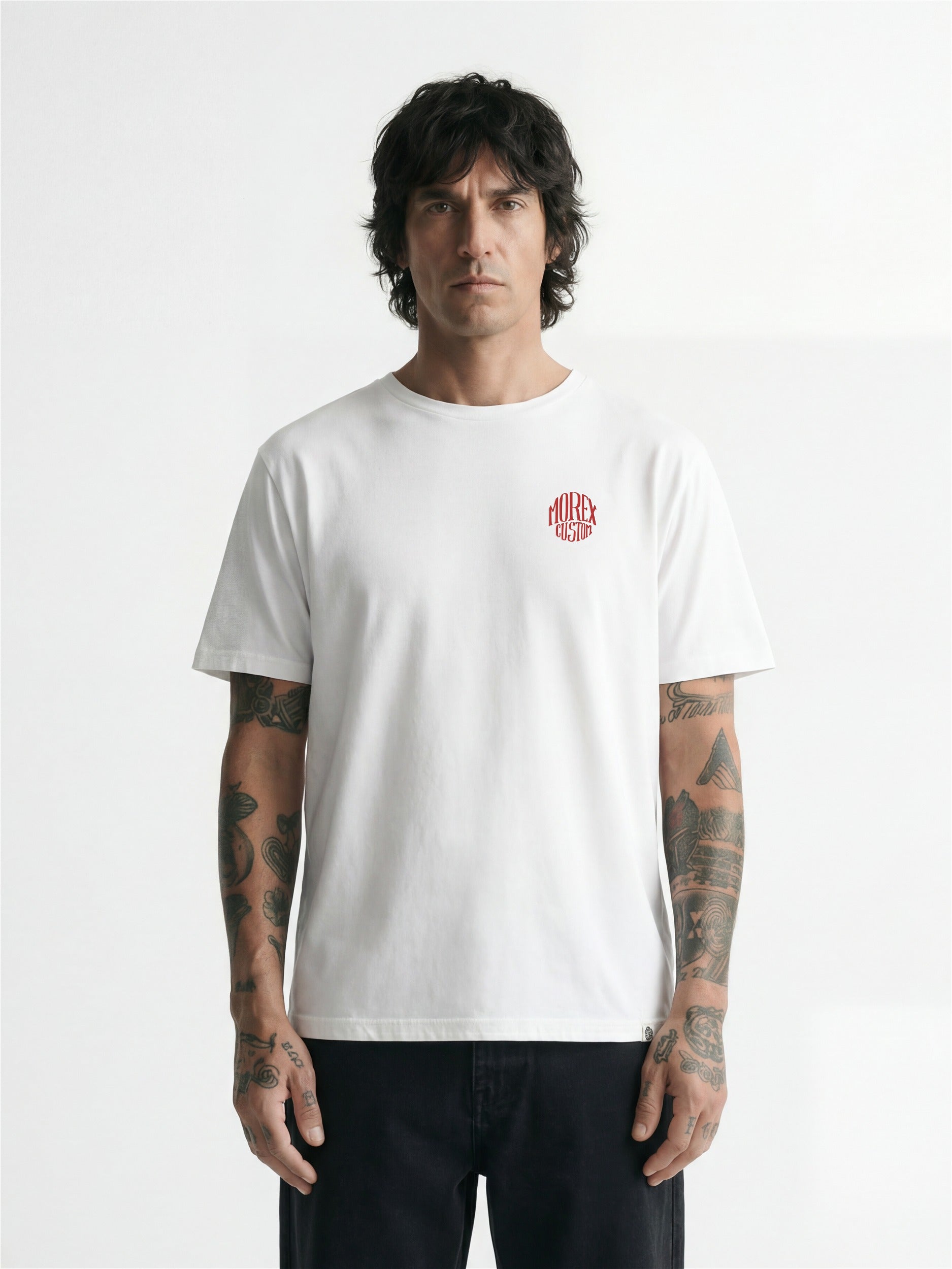 Homme tatoué portant un t-shirt blanc à manches courtes Morex Custom avec un logo circulaire rouge sur la poitrine, style lifestyle décontracté.
