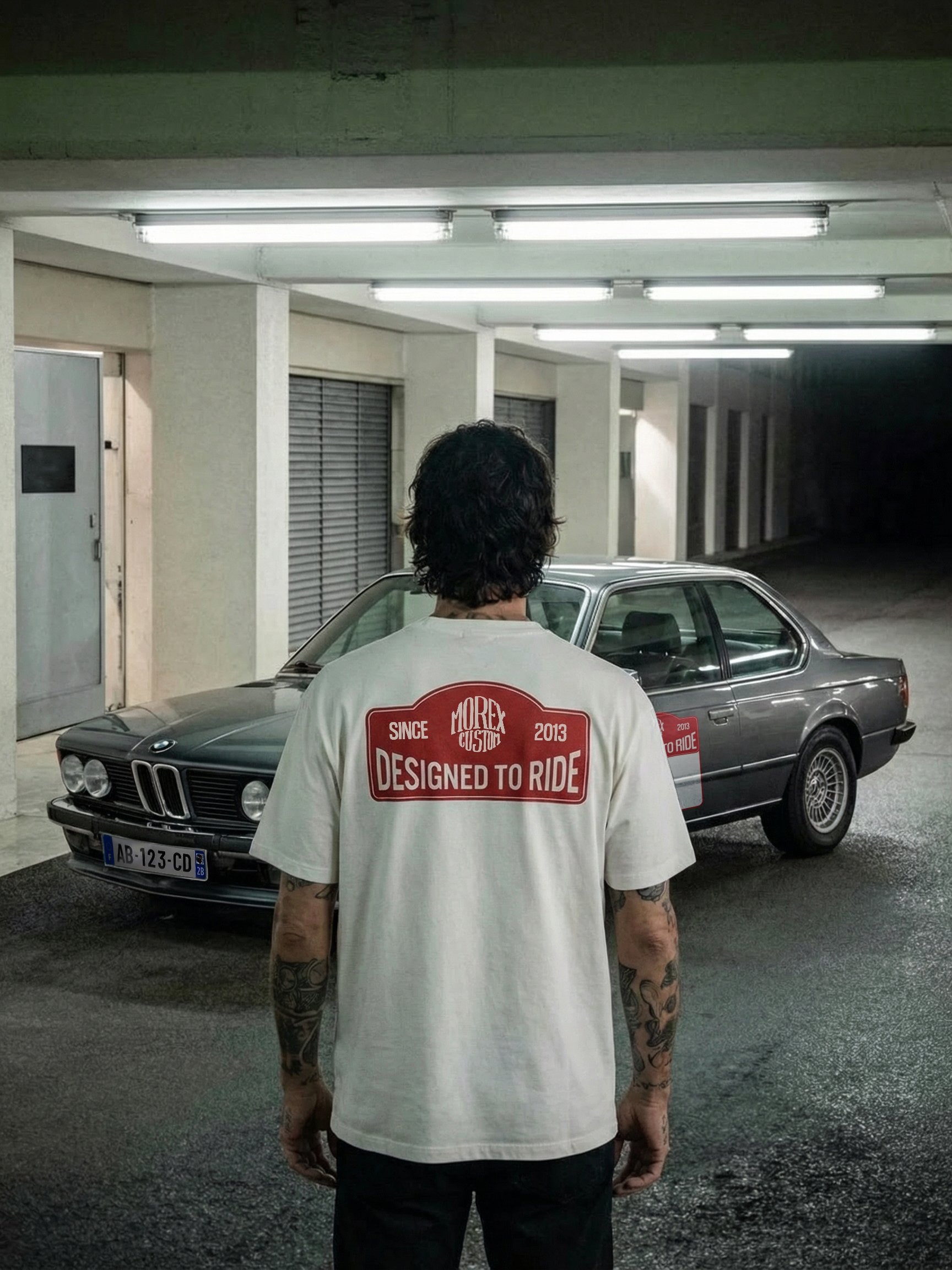 Homme portant un t-shirt blanc Morex Custom avec l'inscription 'Designed to Ride' de dos, devant une BMW de collection grise dans un parking souterrain.
