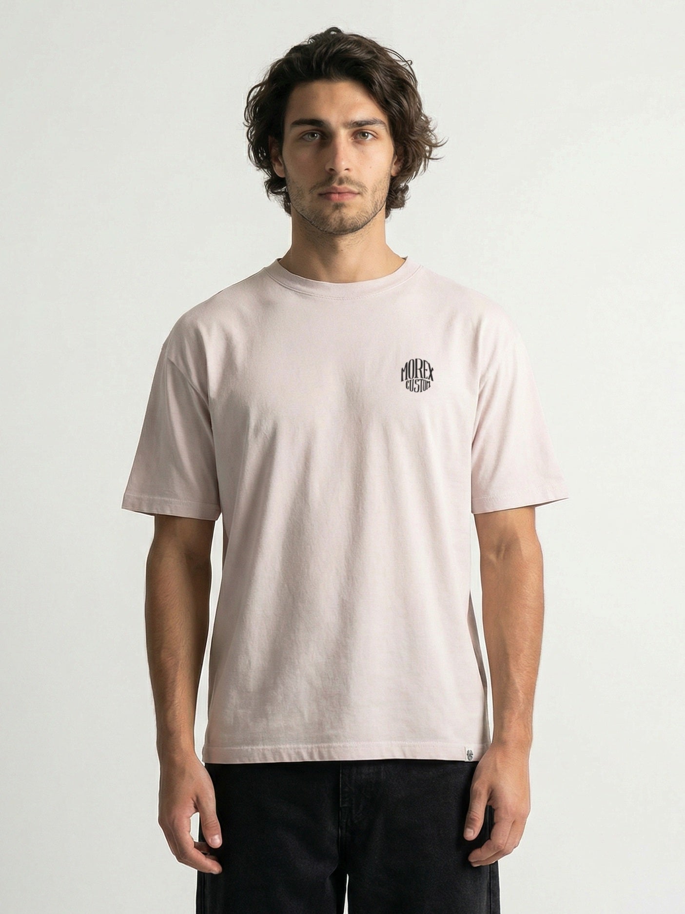 Un homme portant un t-shirt Morex Custom de couleur rose pâle avec un petit logo circulaire sur la poitrine, style lifestyle décontracté sur fond blanc.