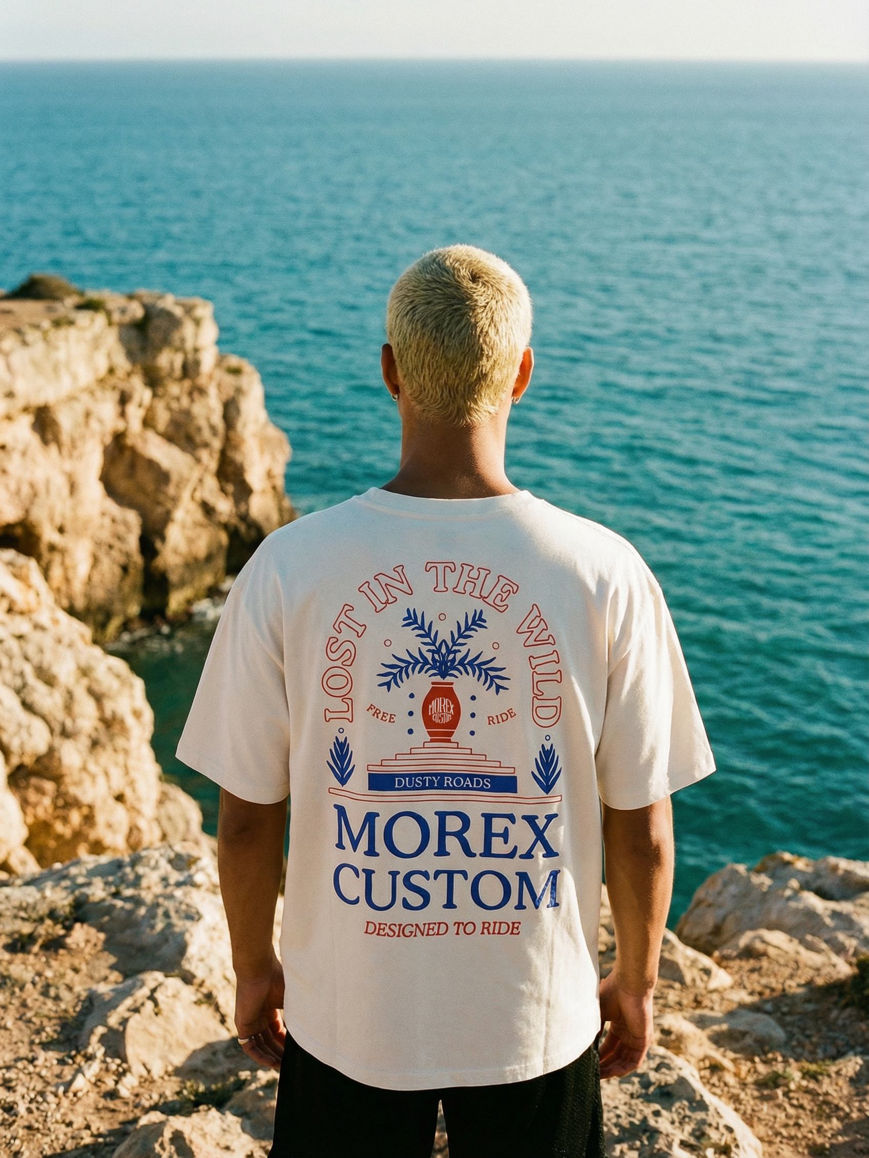 Un homme de dos portant un t-shirt blanc Morex Custom avec le graphique Lost in the Wild devant un paysage côtier et la mer.