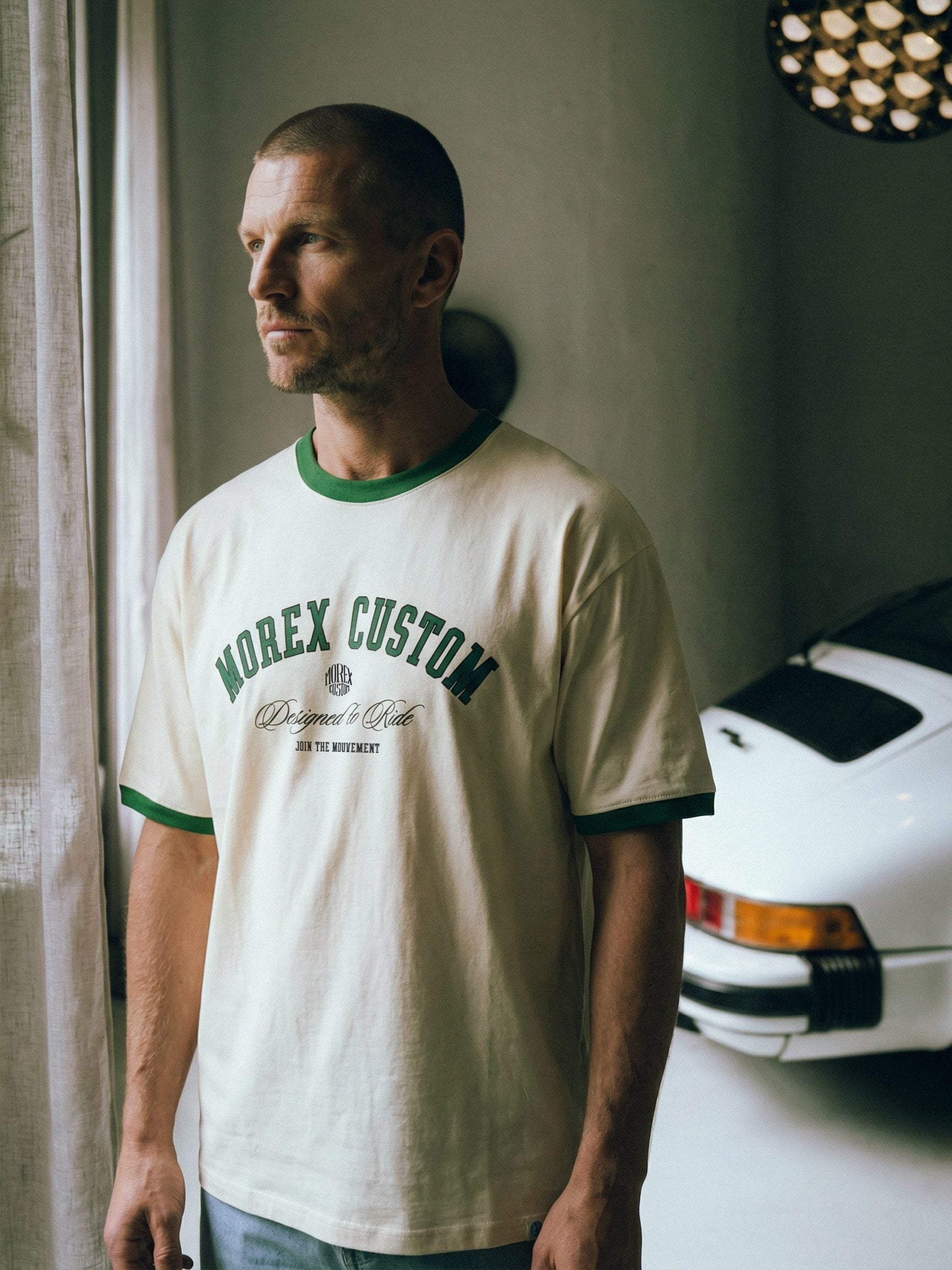 Un homme portant un t-shirt Morex Custom blanc cassé avec des bords verts, posant à côté d'une voiture Porsche classique blanche dans un intérieur de style industriel.