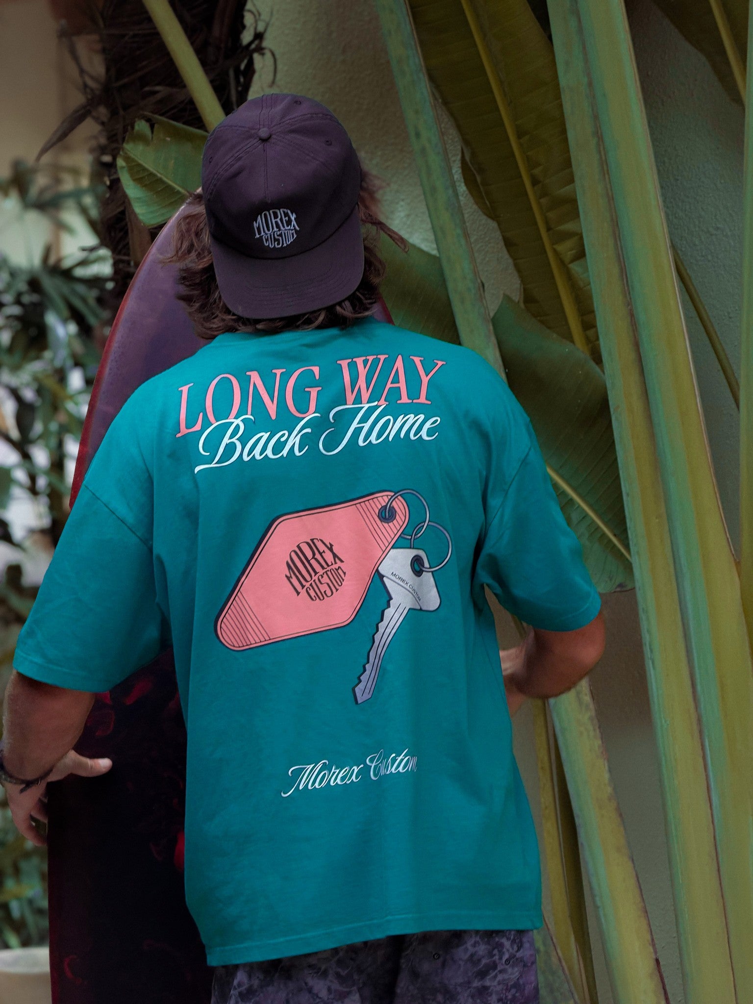 Un homme de dos portant un t-shirt turquoise Morex Custom avec le graphisme Long Way Back Home et une illustration de clé vintage, style lifestyle et surf.