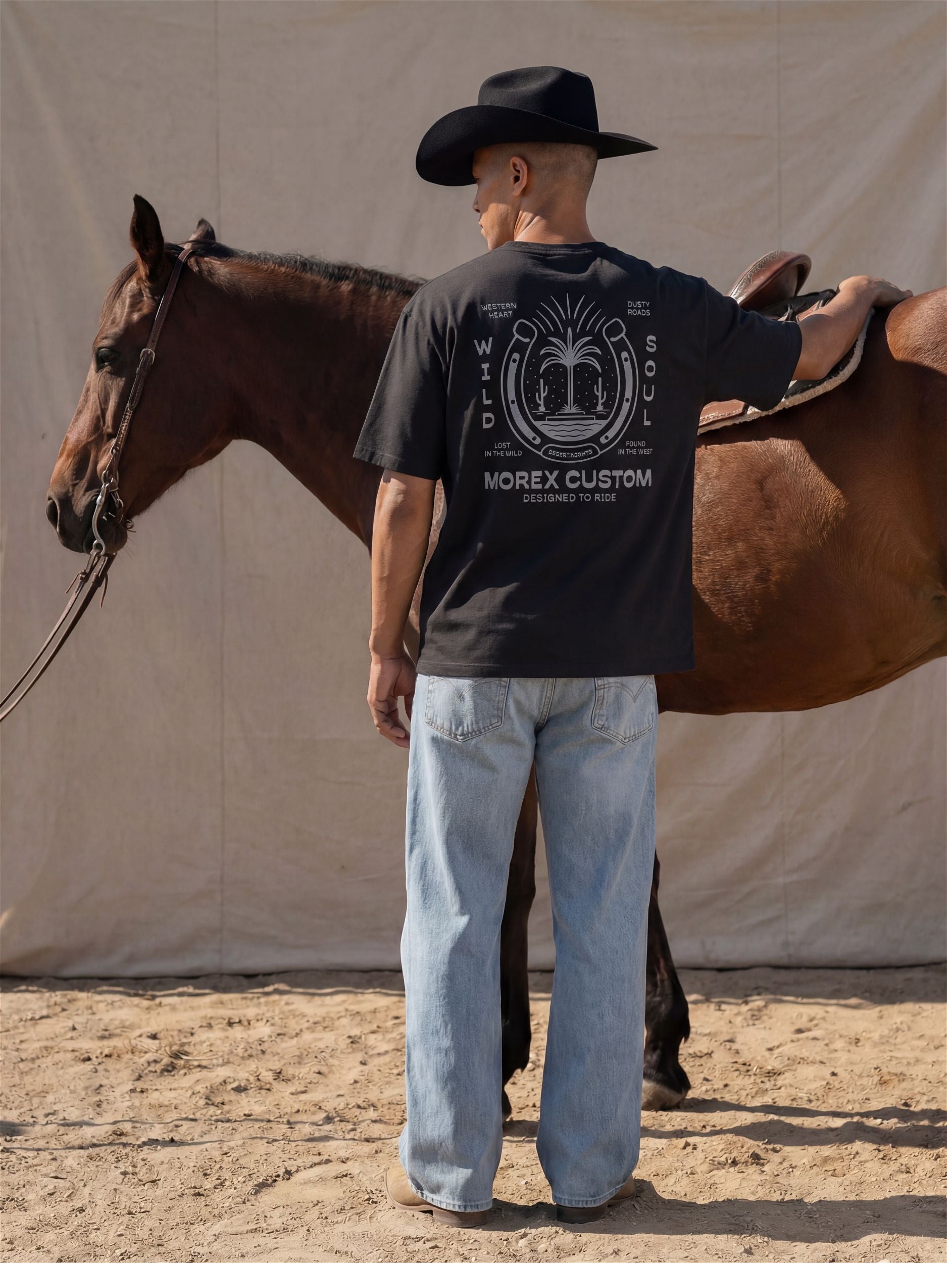 Homme de dos portant un t-shirt noir Morex Custom Wild Soul et un chapeau de cowboy, debout à côté d'un cheval marron.