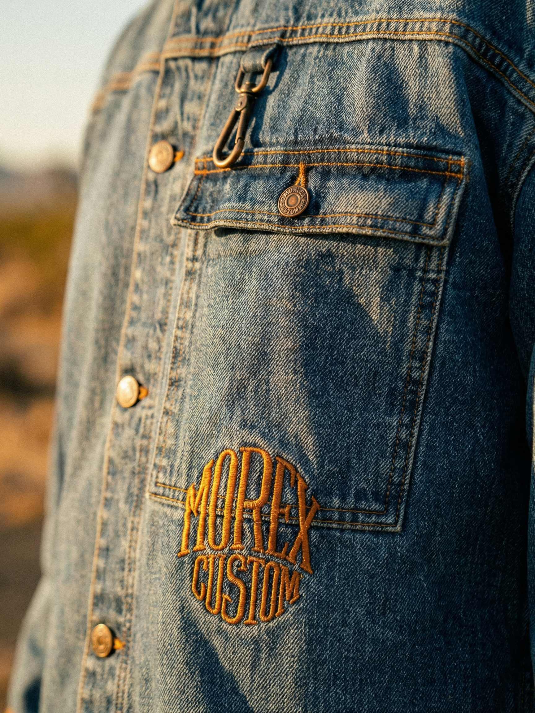 Gros plan sur une veste en jean Morex Custom arborant une broderie circulaire orange et un crochet métallique, style lifestyle et moto.