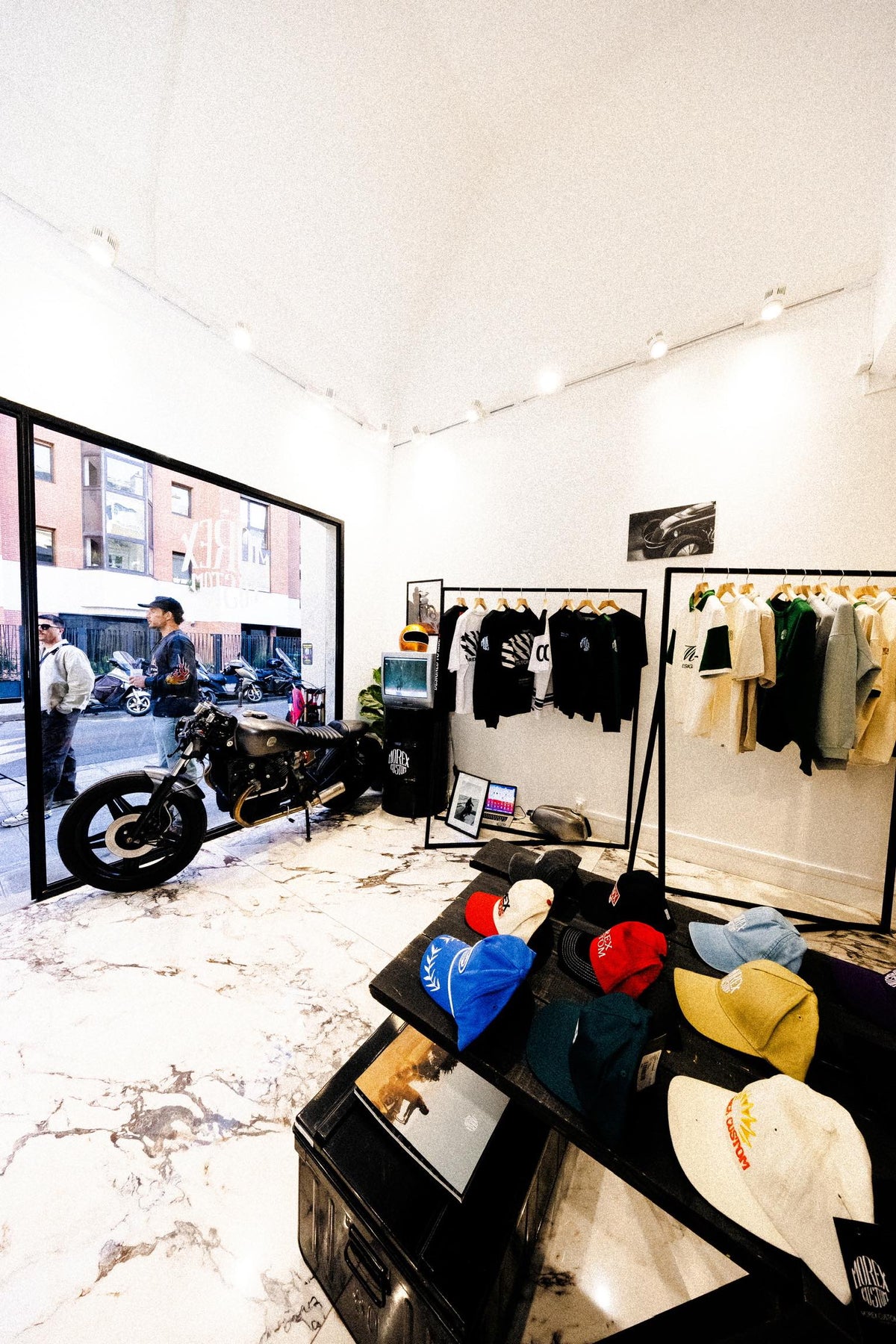 Vue intérieure du showroom Morex Custom avec une moto de collection exposée et une sélection de vêtements lifestyle, t-shirts et casquettes.