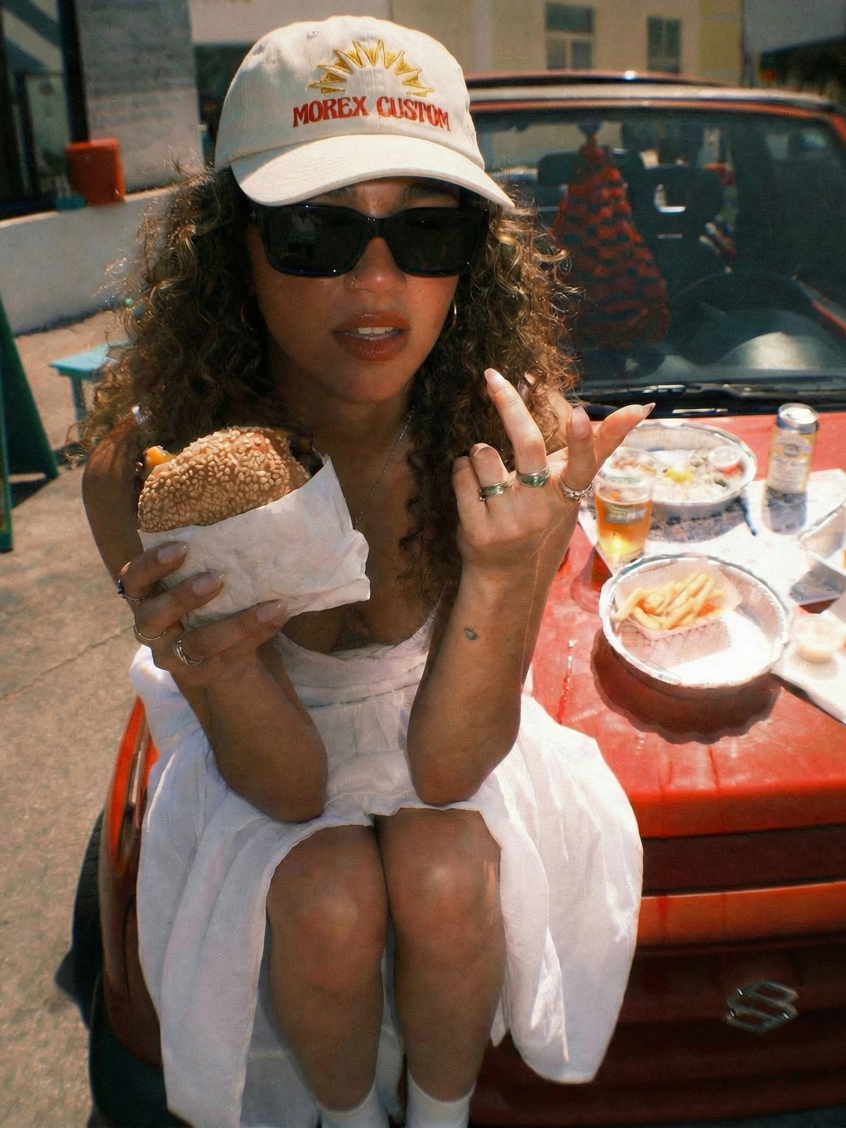 Femme au style urbain décontracté portant une casquette blanche brodée Morex Custom, mangeant un burger sur le capot d'une voiture rouge, illustrant l'univers lifestyle de la marque.