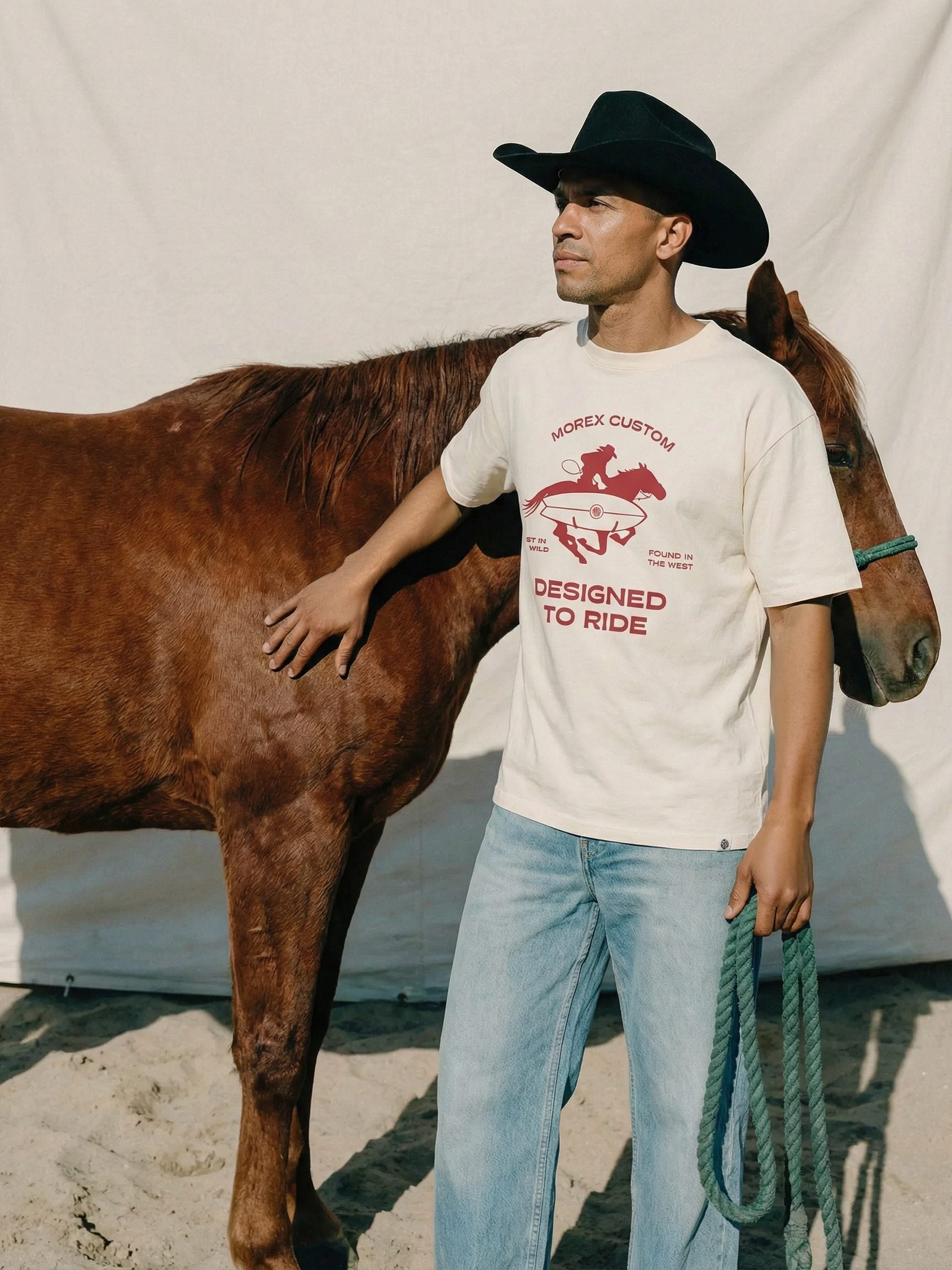 Man in a black cowboy hat wearing a Morex Custom 'Designed to Ride' white graphic t-shirt standing next to a brown horse.