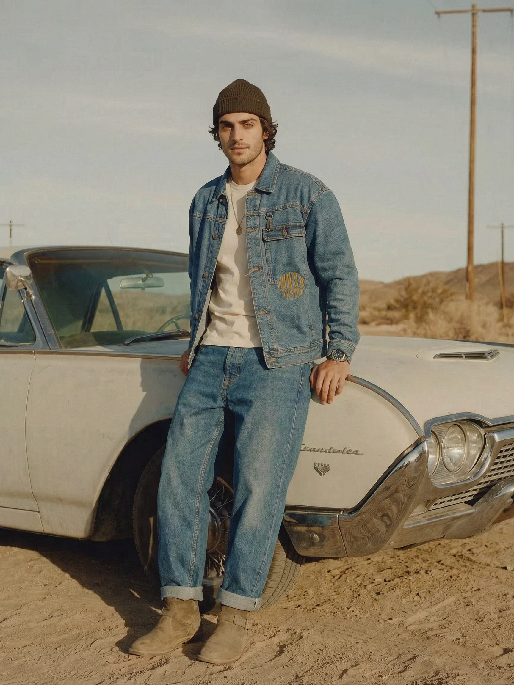 Un homme portant une veste en jean et un bonnet Morex Custom, s'appuyant contre une voiture vintage blanche dans un décor de désert.
