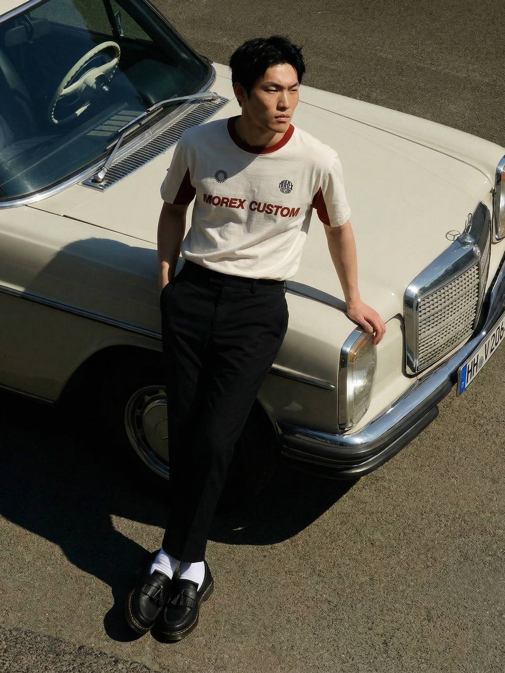 Homme portant un t-shirt Morex Custom blanc cassé et bordeaux avec un pantalon noir, s'appuyant contre le capot d'une Mercedes vintage beige.