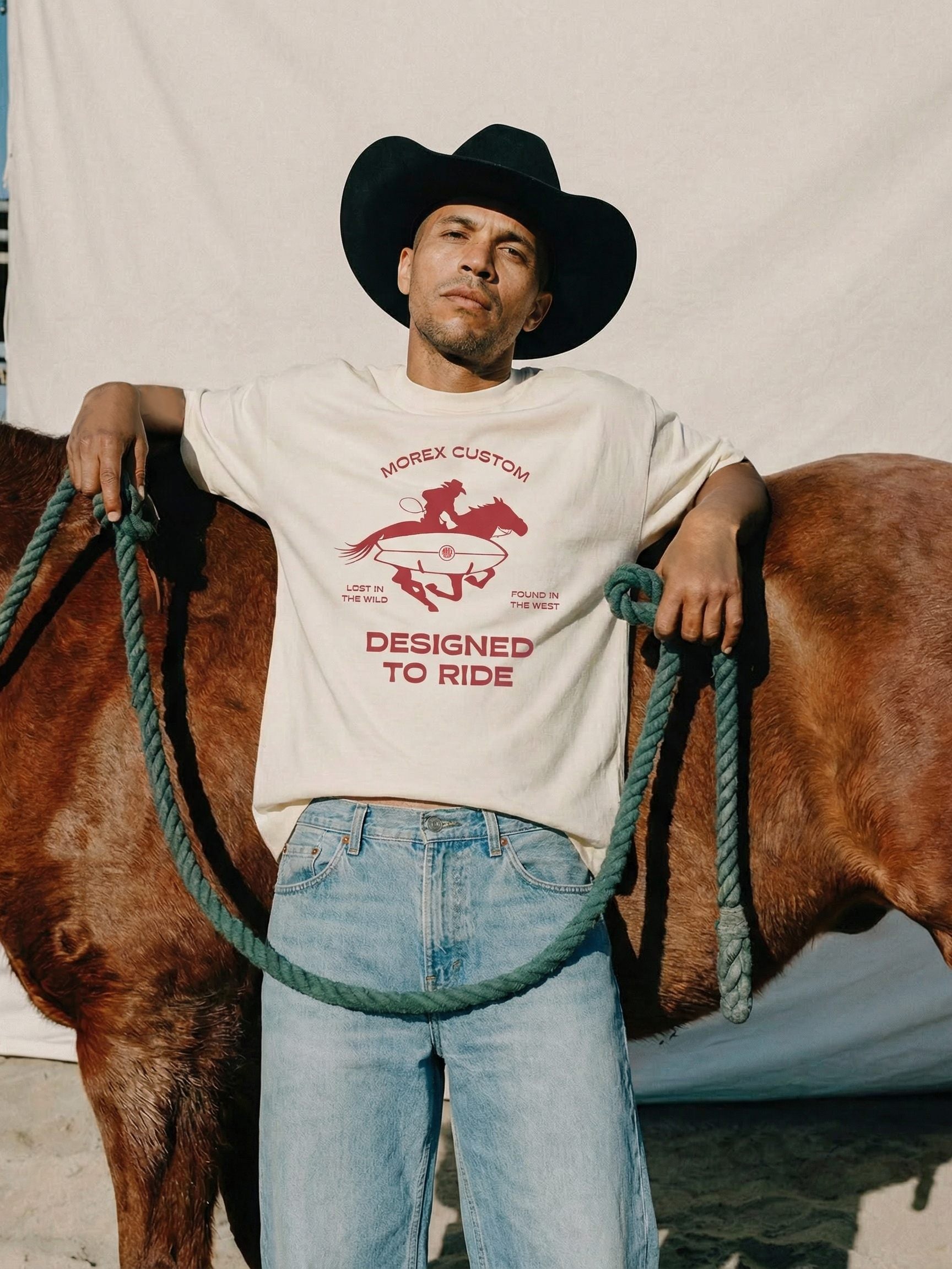 Un homme portant un chapeau de cowboy et un t-shirt blanc Morex Custom 'Designed to Ride' s'appuyant sur un cheval brun.