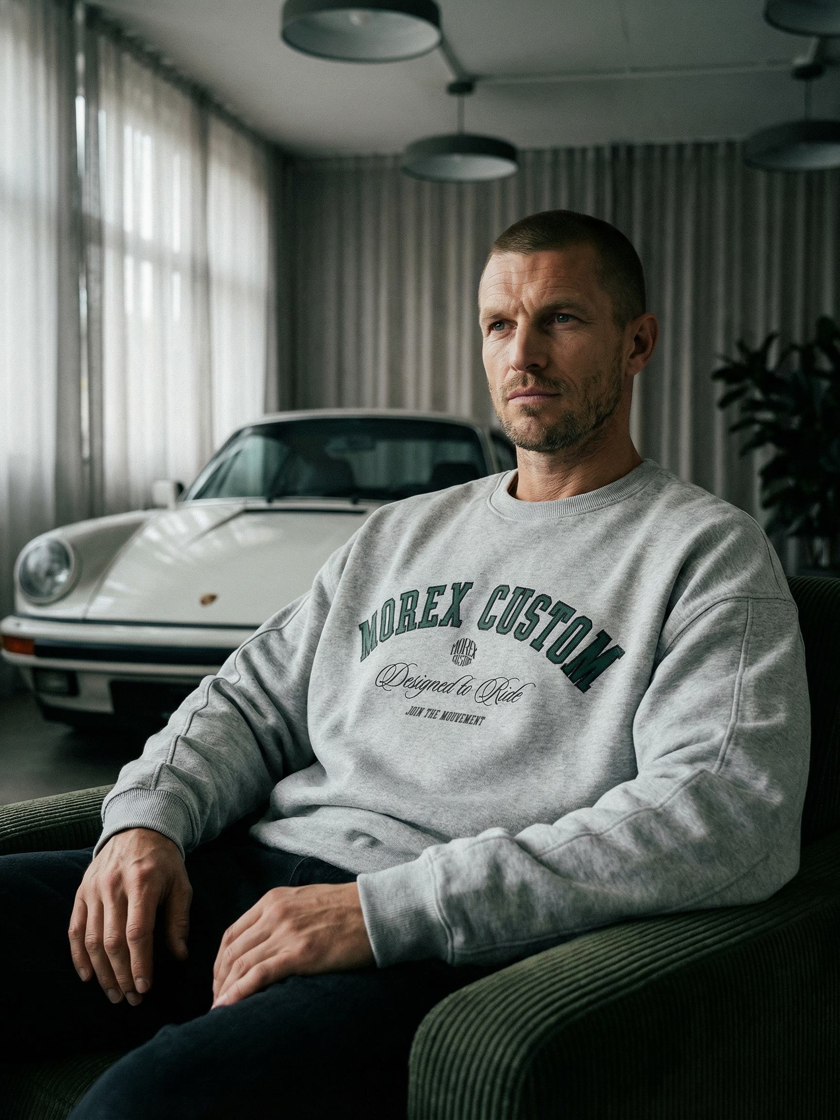 Homme portant un sweatshirt gris Morex Custom assis devant une Porsche classique blanche dans un intérieur moderne.