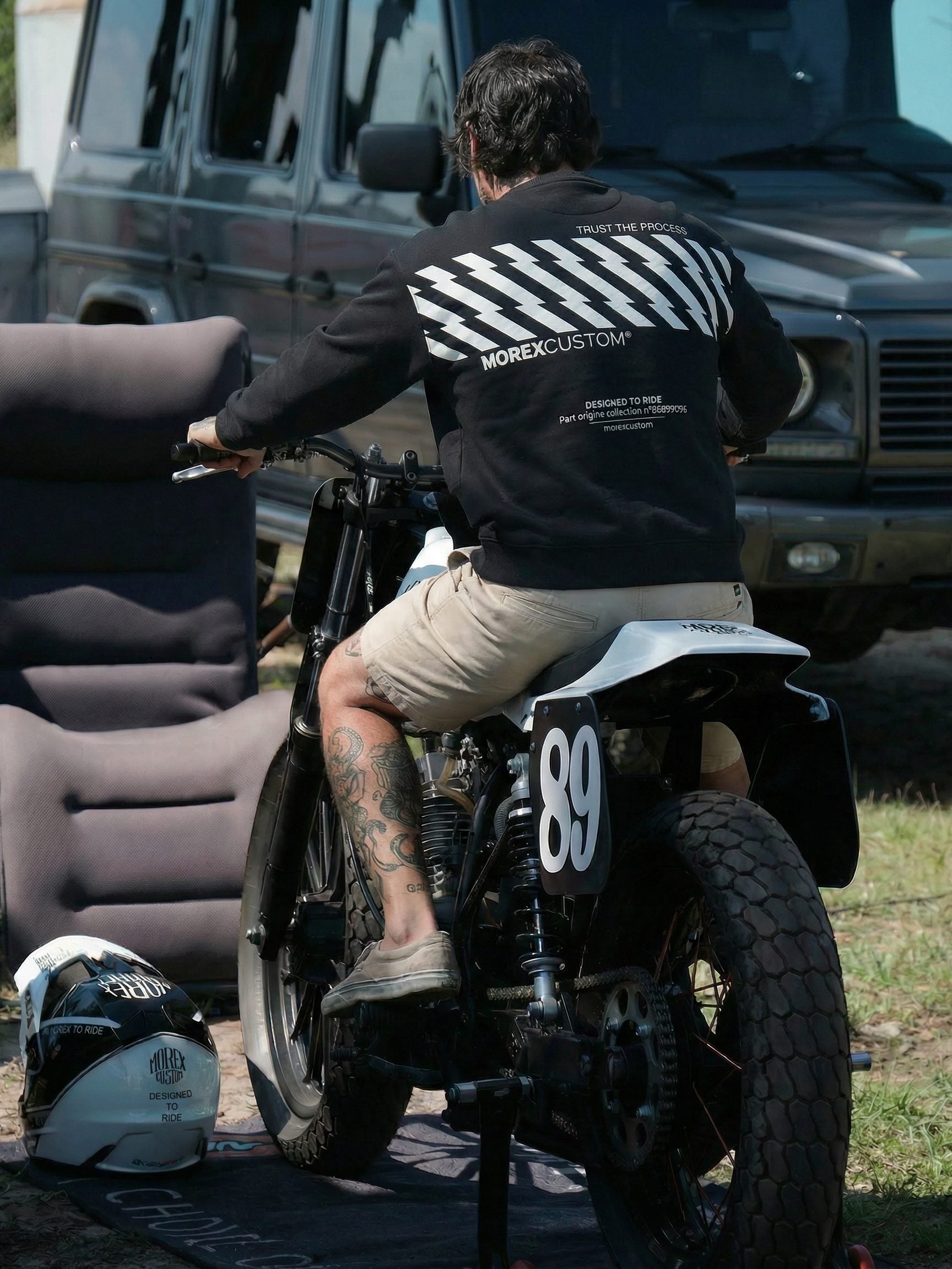 Un homme de dos portant un sweat-shirt Morex Custom noir avec des motifs graphiques blancs, assis sur une moto de customisation numéro 89.