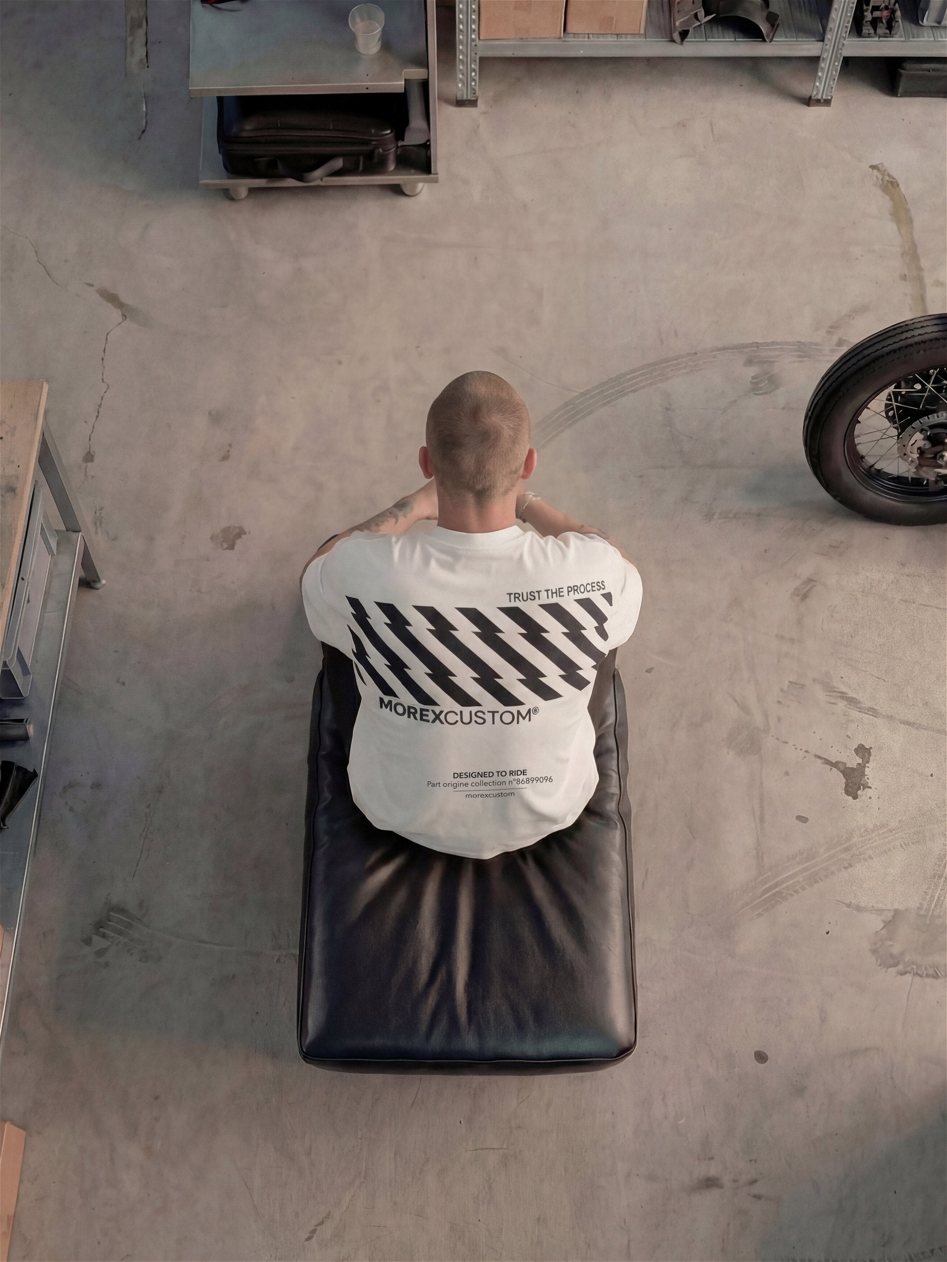 Vue de dessus d'un homme portant un t-shirt blanc de la marque Morex Custom avec des motifs graphiques noirs, assis sur un siège en cuir noir au milieu d'un atelier de moto.