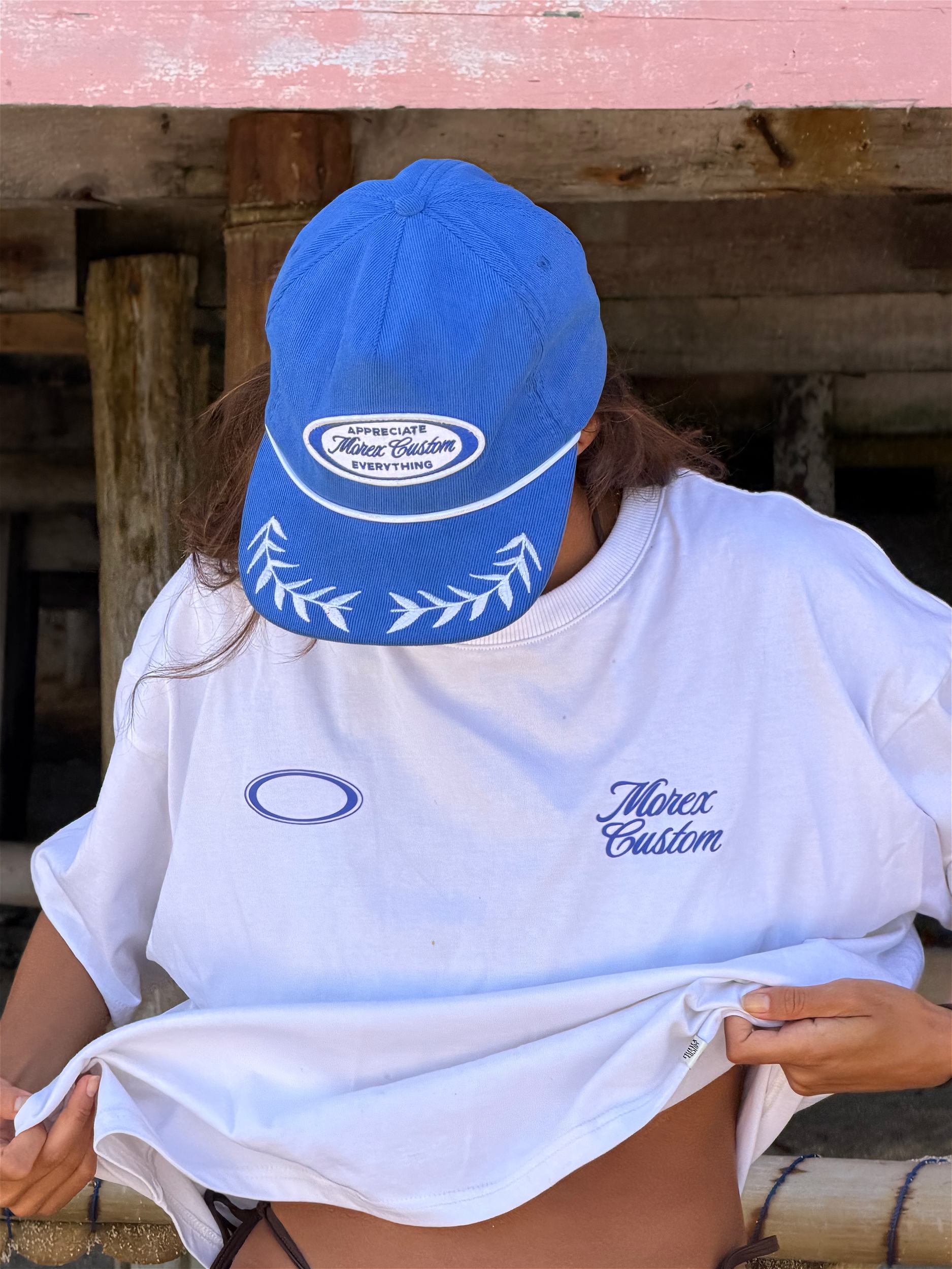 Femme portant un t-shirt blanc Morex Custom et une casquette bleue logotypée dans un style lifestyle de bord de mer.