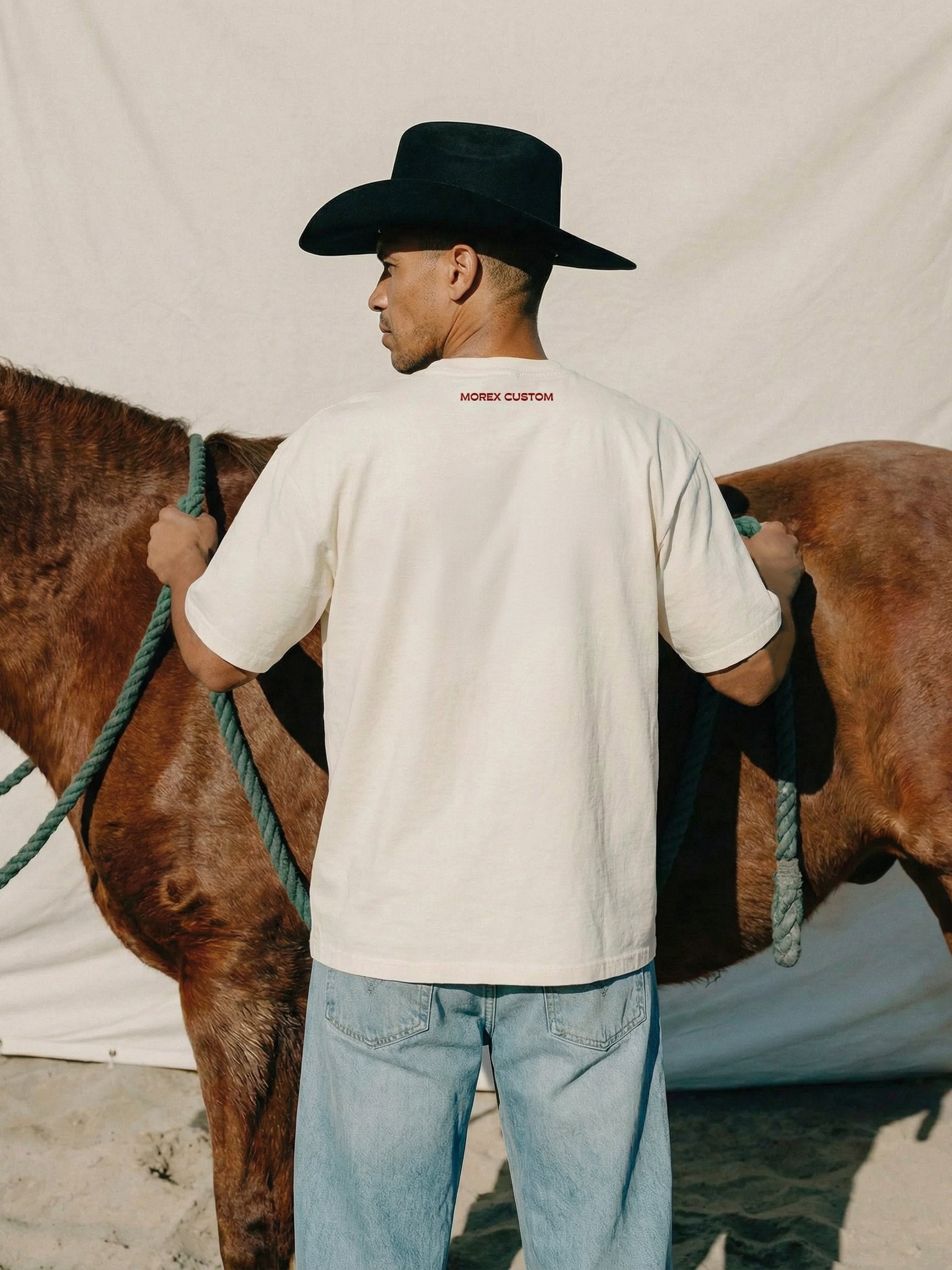 Homme de dos portant un t-shirt blanc Morex Custom et un chapeau de cowboy noir, tenant un cheval marron dans un décor lifestyle rustique.
