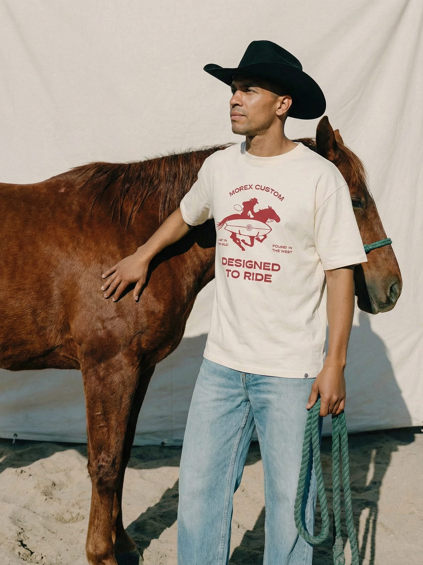Un homme portant un t-shirt graphique Morex Custom 'Designed to Ride' et un chapeau de cowboy noir, posant à côté d'un cheval brun.