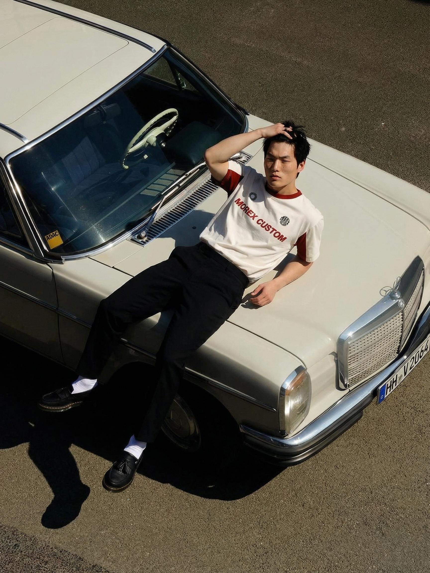 Un homme portant un t-shirt blanc Morex Custom avec des détails rouges, assis sur le capot d'une Mercedes vintage blanche dans un style lifestyle urbain.
