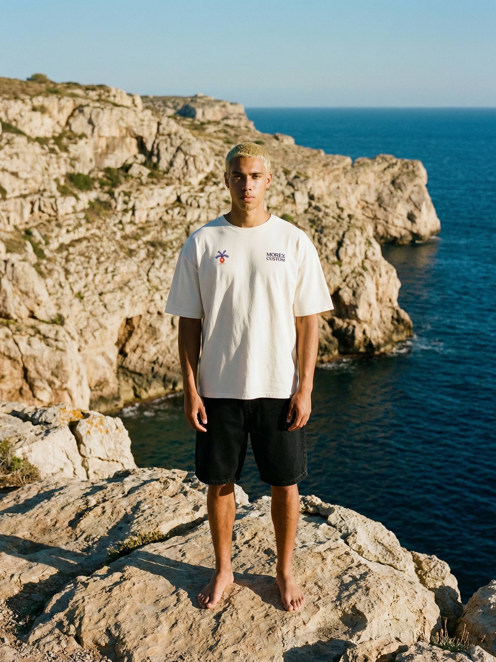 Un homme portant un t-shirt blanc Morex Custom et un short noir, debout sur une falaise rocheuse devant l'océan.
