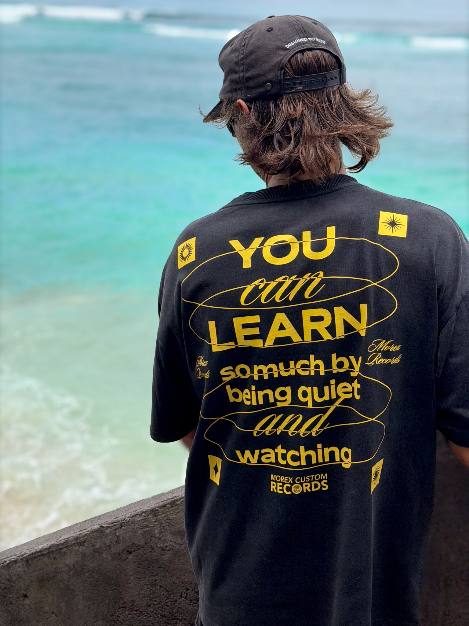 T-shirt noir Morex Custom avec message graphique jaune 'You can learn so much by being quiet and watching' porté par un homme face à la mer.