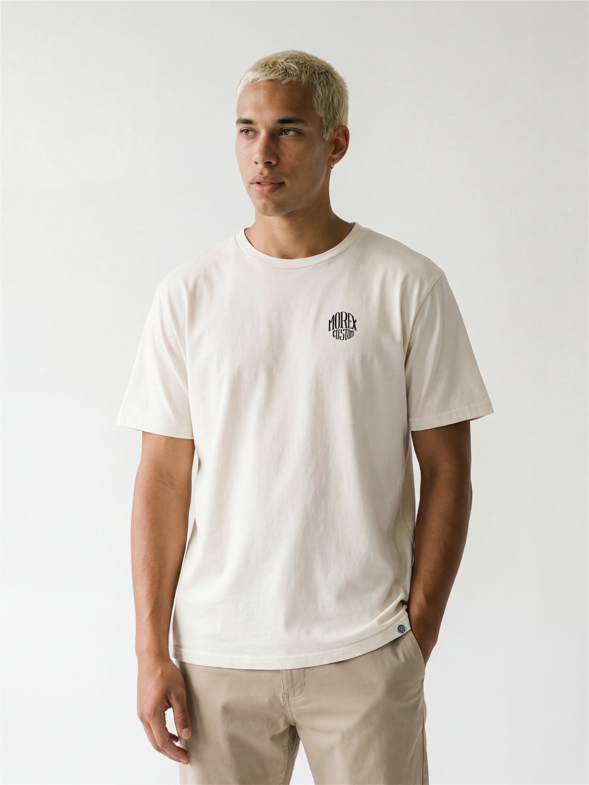 Homme portant un tee-shirt blanc cassé de la marque Morex Custom avec un petit logo circulaire sur la poitrine, style décontracté et moto.