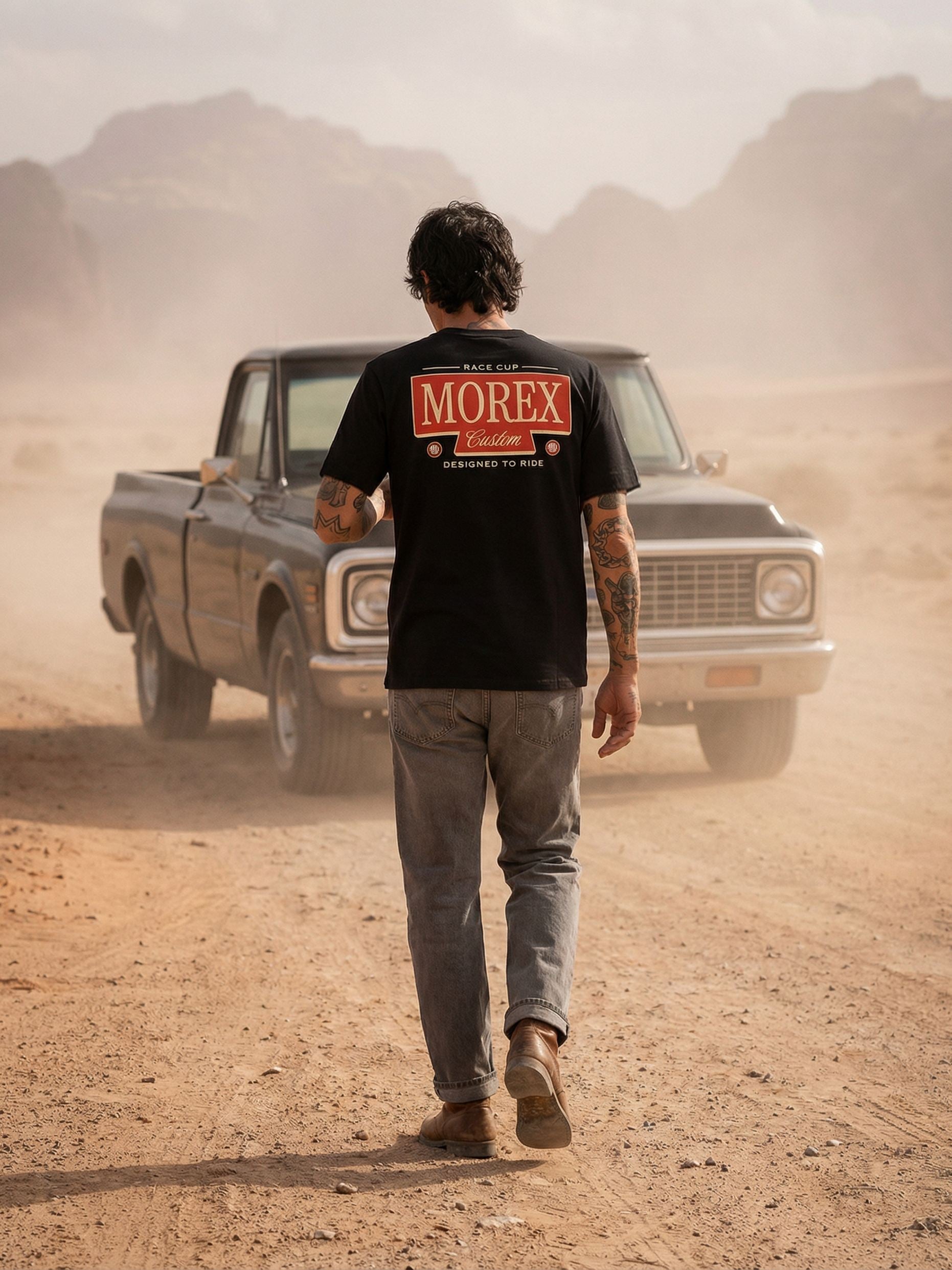 Homme de dos portant un t-shirt noir Morex Custom marchant vers un vieux pick-up dans un décor désertique poussiéreux.