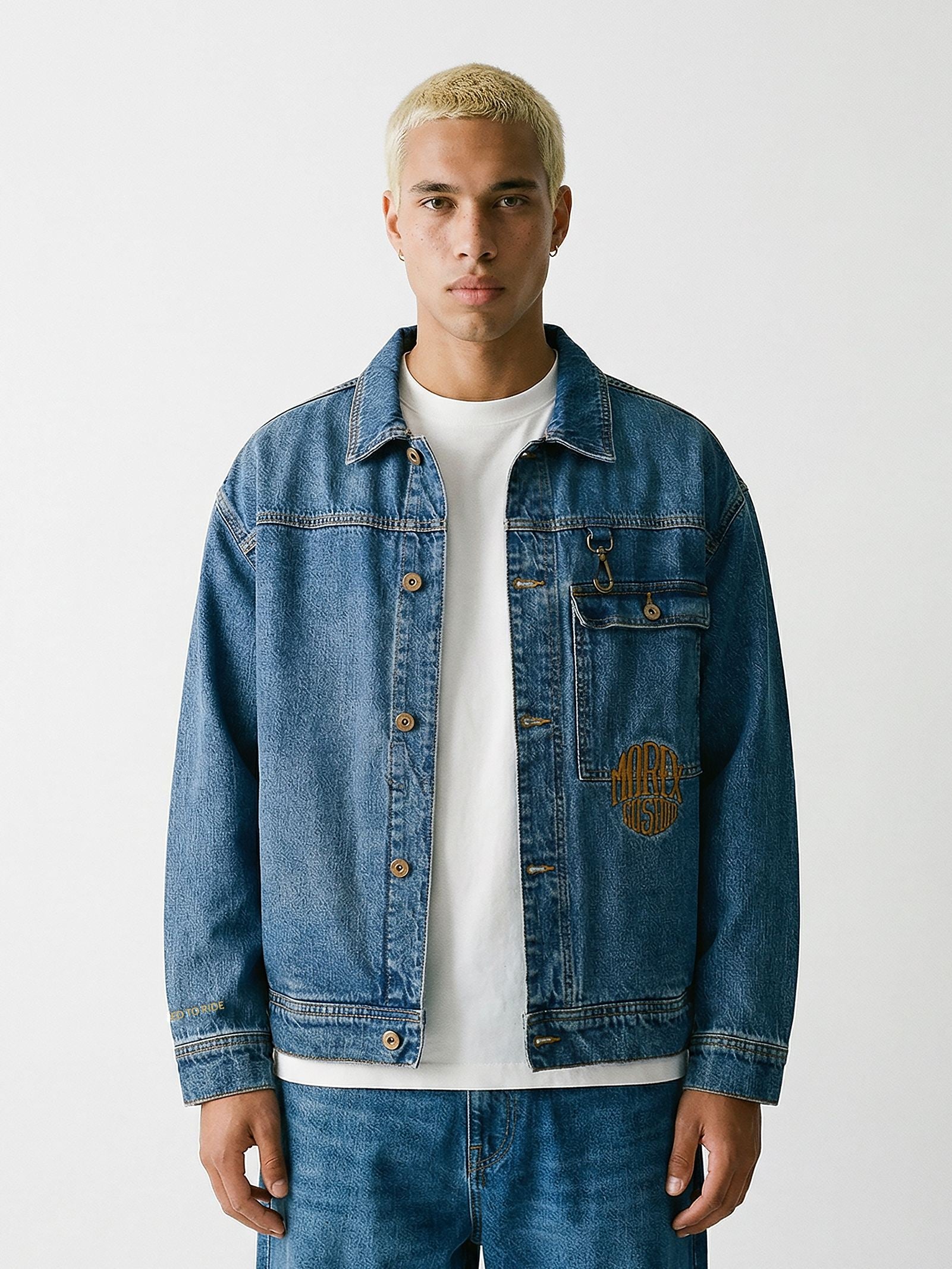 Un homme aux cheveux courts et décolorés portant une veste en jean bleu délavé de la marque Morex Custom, sur un t-shirt blanc et un pantalon assorti.