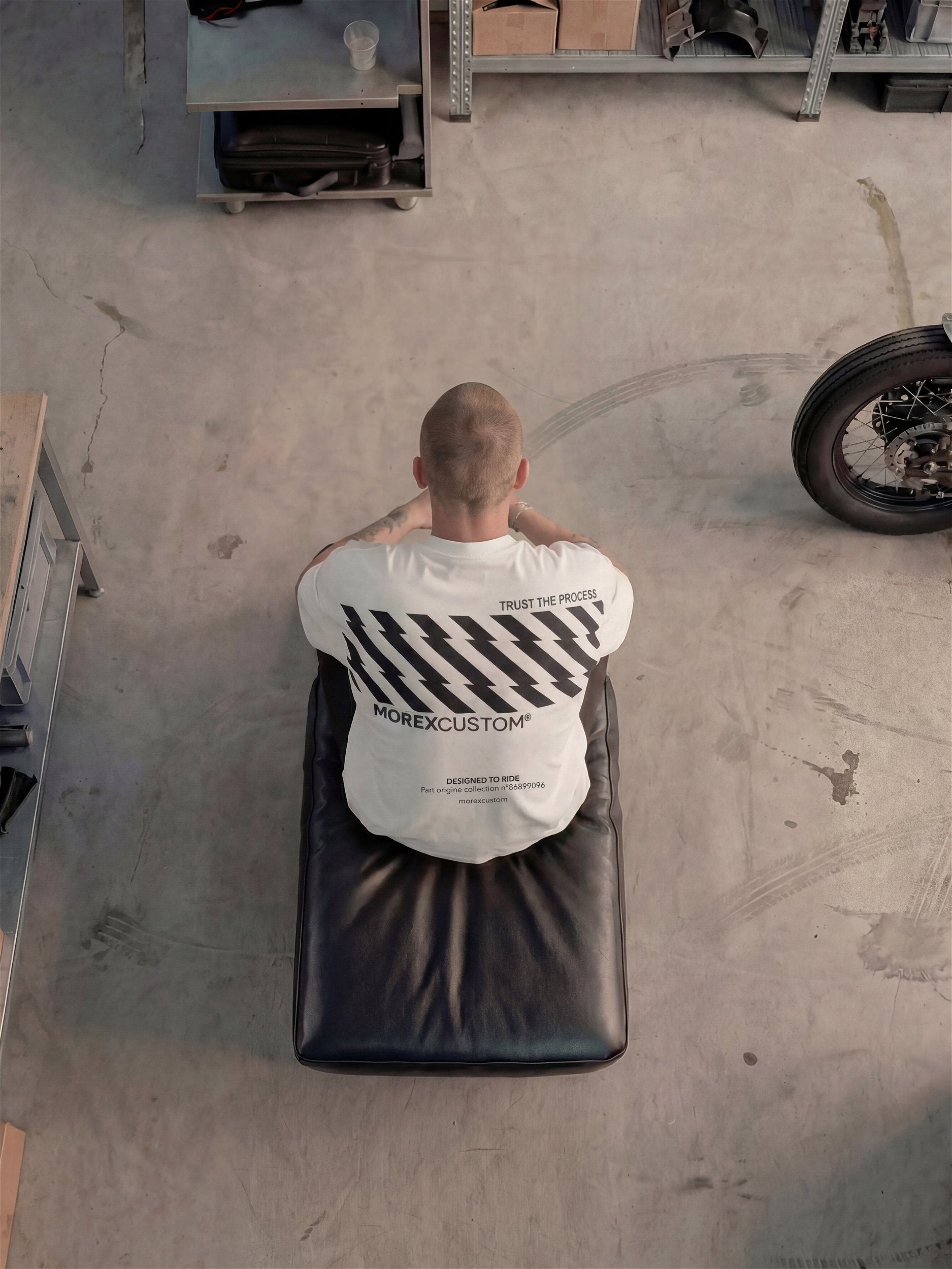 Une vue plongeante d'un homme assis sur un siège en cuir noir dans un atelier, portant un t-shirt Morex Custom blanc arborant des rayures noires diagonales et le logo de la marque. En arrière-plan, on aperçoit des étagères de rangement remplies de boîtes et d'outils, ainsi que la roue arrière d'une moto personnalisée sur le sol en béton.