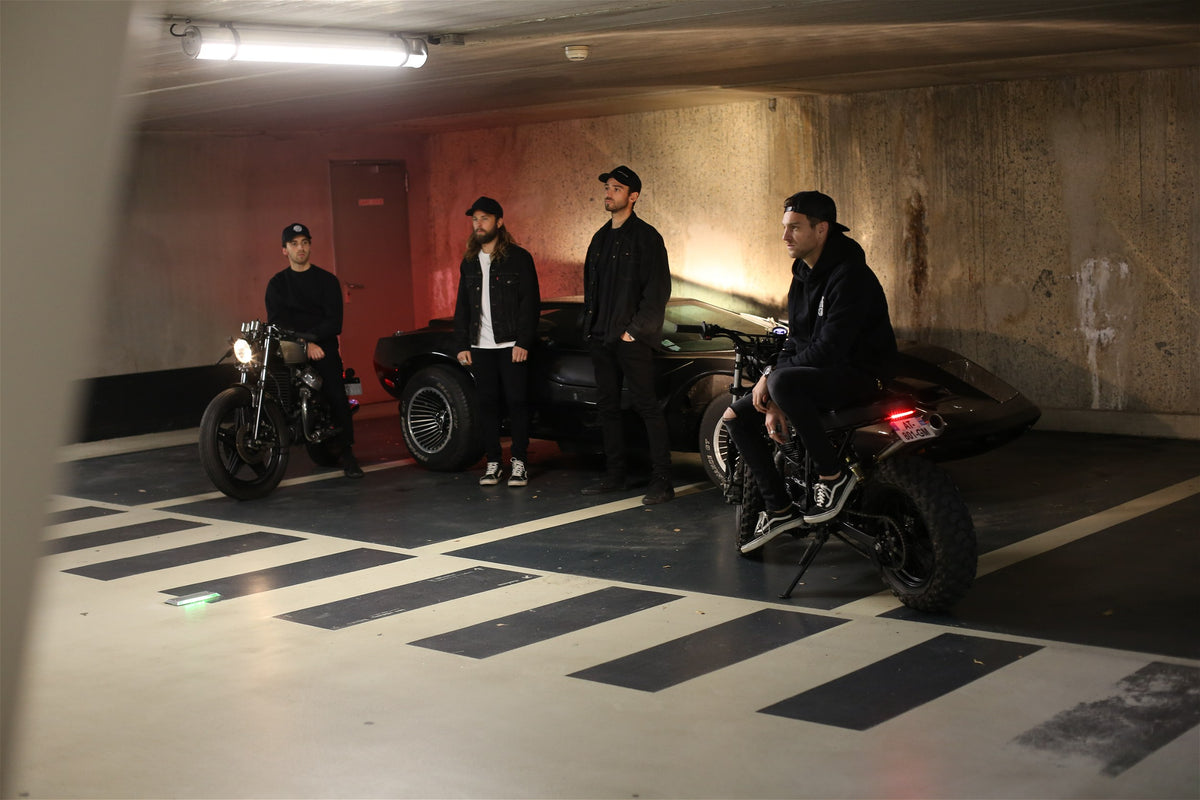 L'équipe Morex Custom posant avec des motos customisées et un véhicule de collection dans un parking souterrain au style industriel.