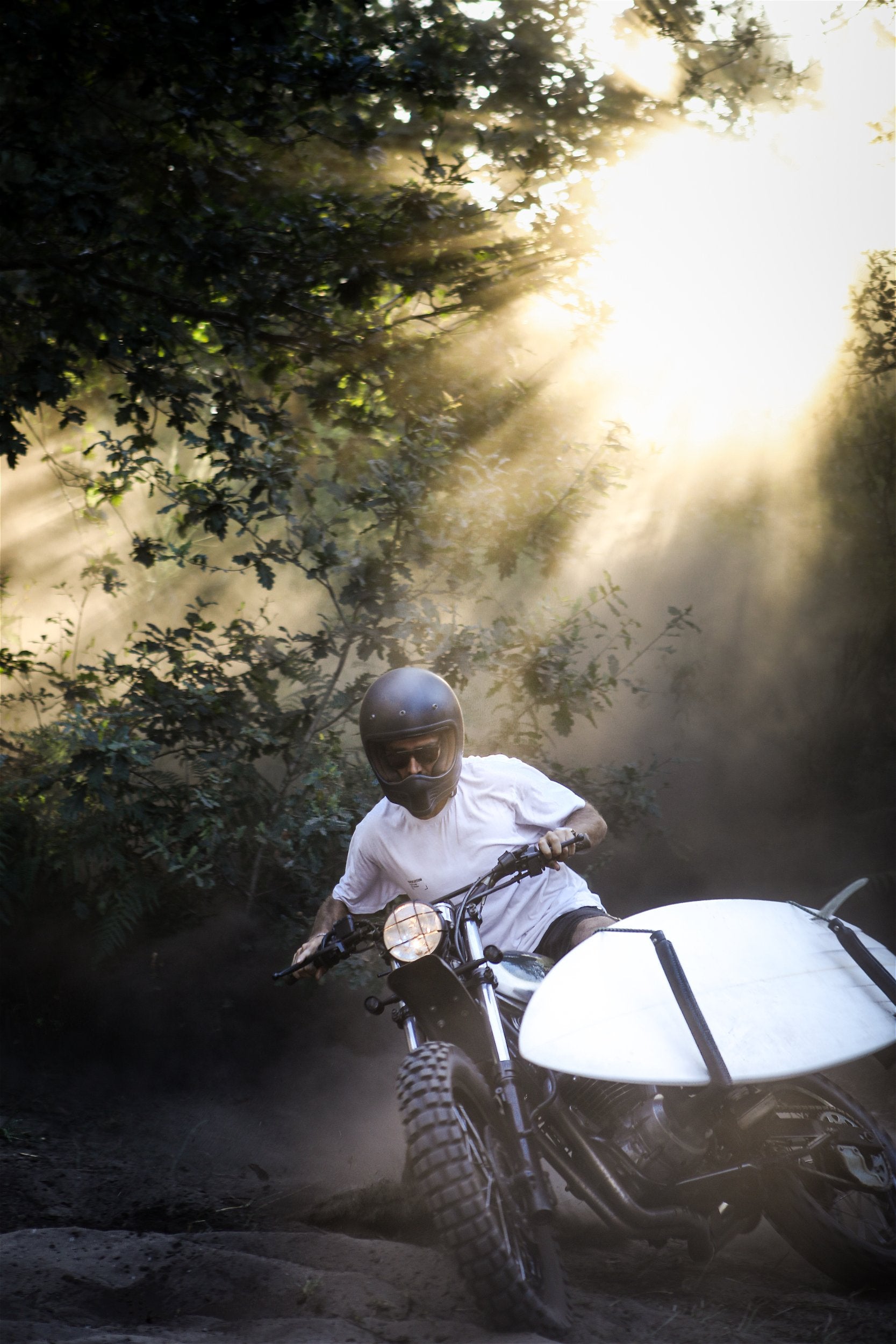 Un pilote sur une moto personnalisée Morex Custom équipée d'un rack de surf, traversant une forêt poussiéreuse sous des rayons de soleil spectaculaires.