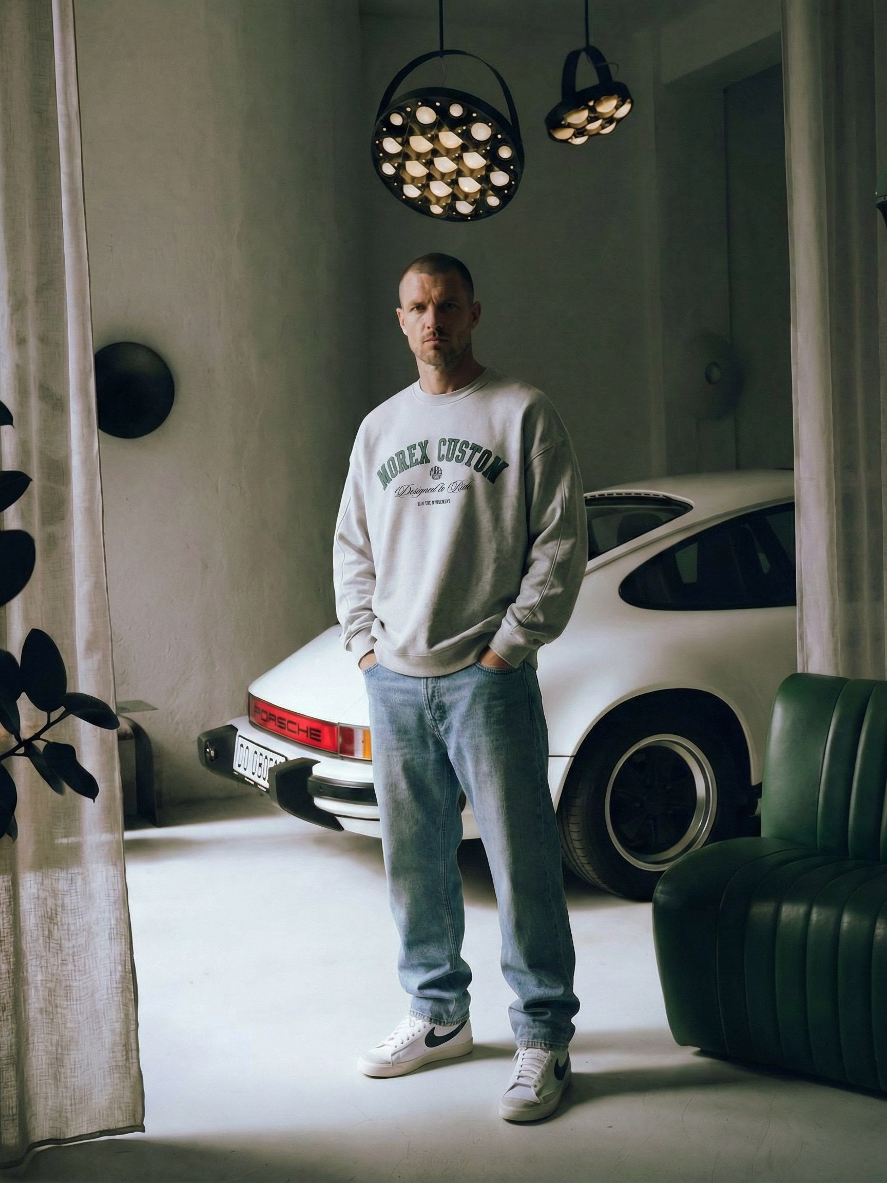Un homme portant un sweatshirt Morex Custom gris et un jean bleu, posant devant une voiture Porsche classique blanche dans un garage au style industriel avec des luminaires design.