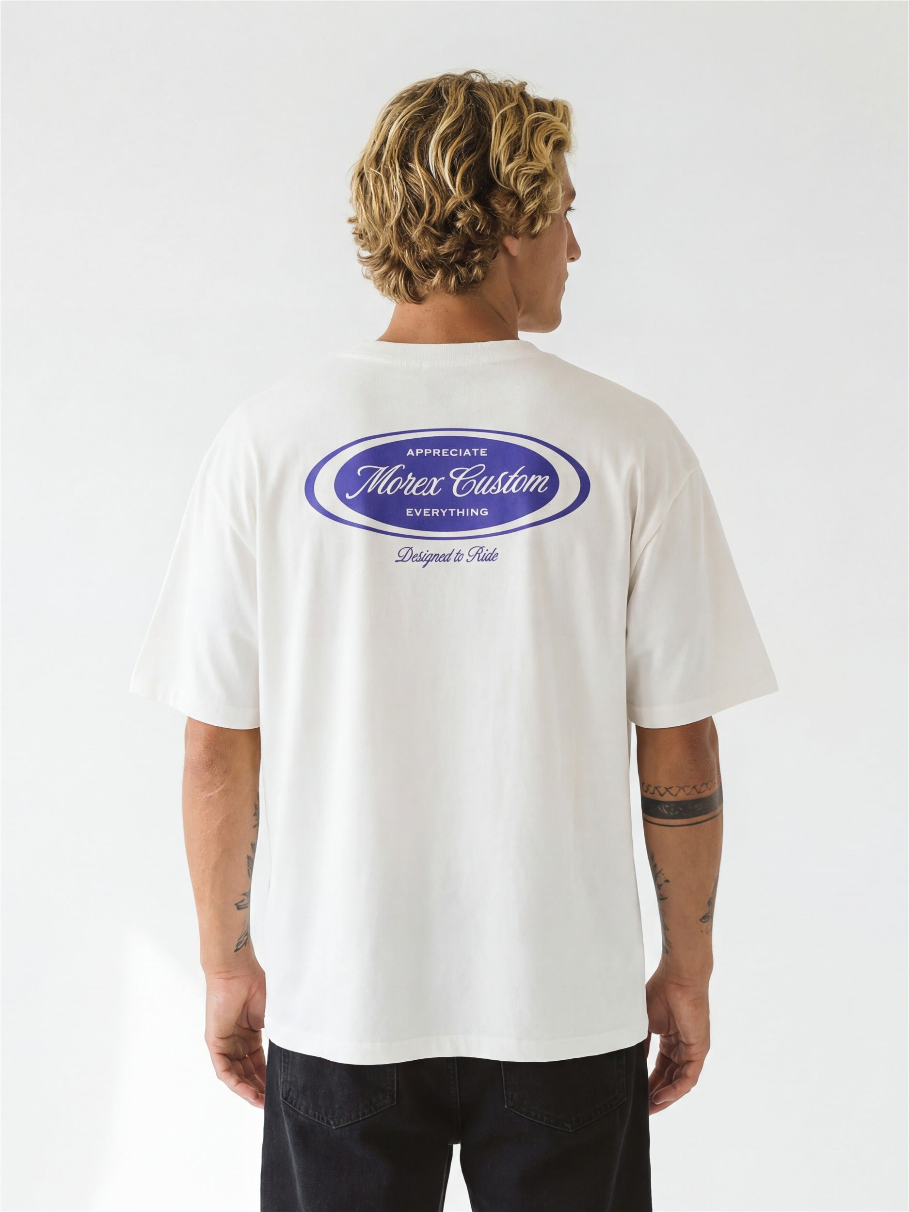 Vue arrière d'un homme portant un t-shirt blanc Morex Custom avec un logo ovale violet 'Morex Custom Designed to Ride' sur le dos.